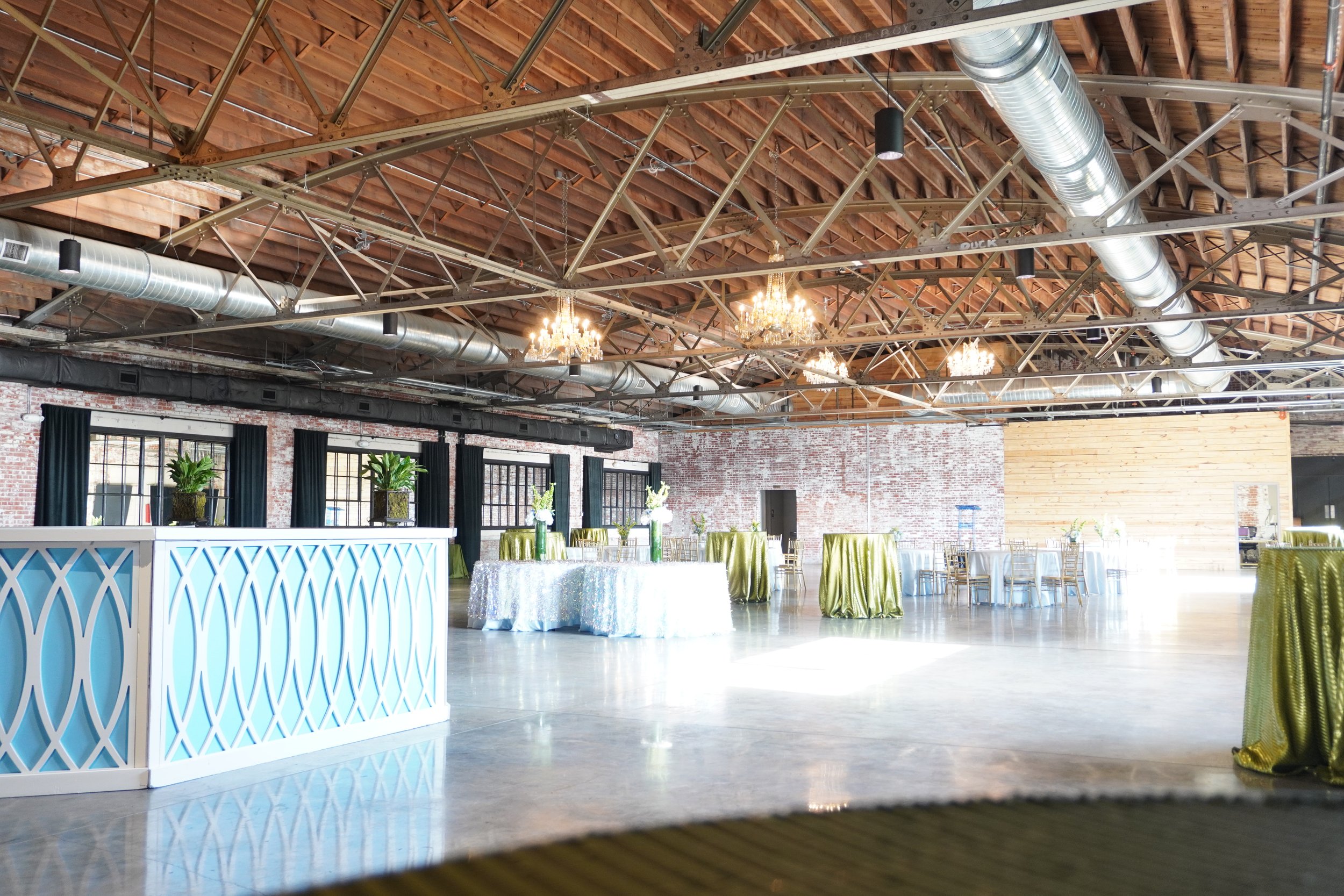 An event space with decorated tables, chandeliers hanging from the wooden ceiling, and large windows with black curtains, in a rustic-industrial style venue.