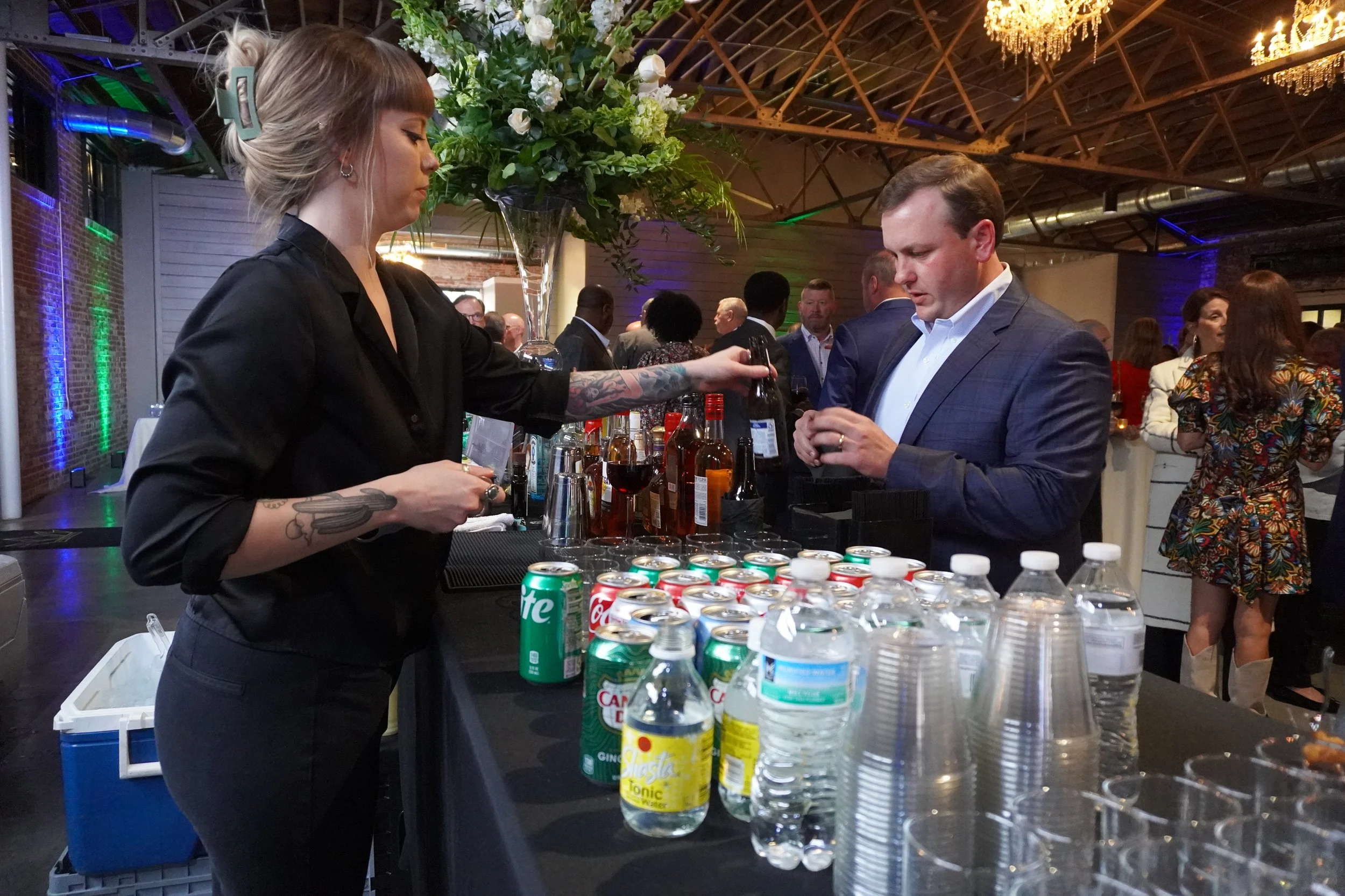 A woman serving drinks at a bar during a social event, with bottles, canned and bottled beverages on the counter and multiple people in the background under a decorated ceiling.
