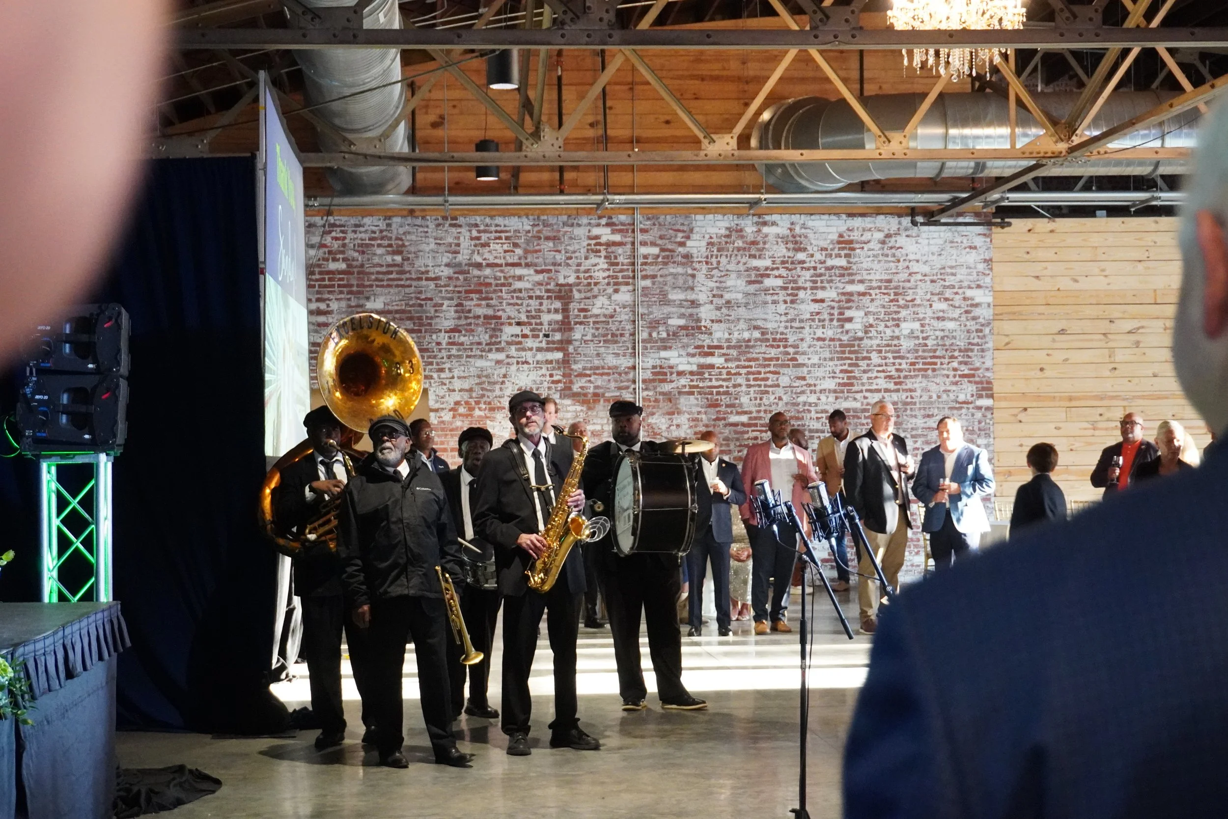 A group of musicians performing on stage at an indoor event, with brick and wood interior walls, and people standing and watching in the background.