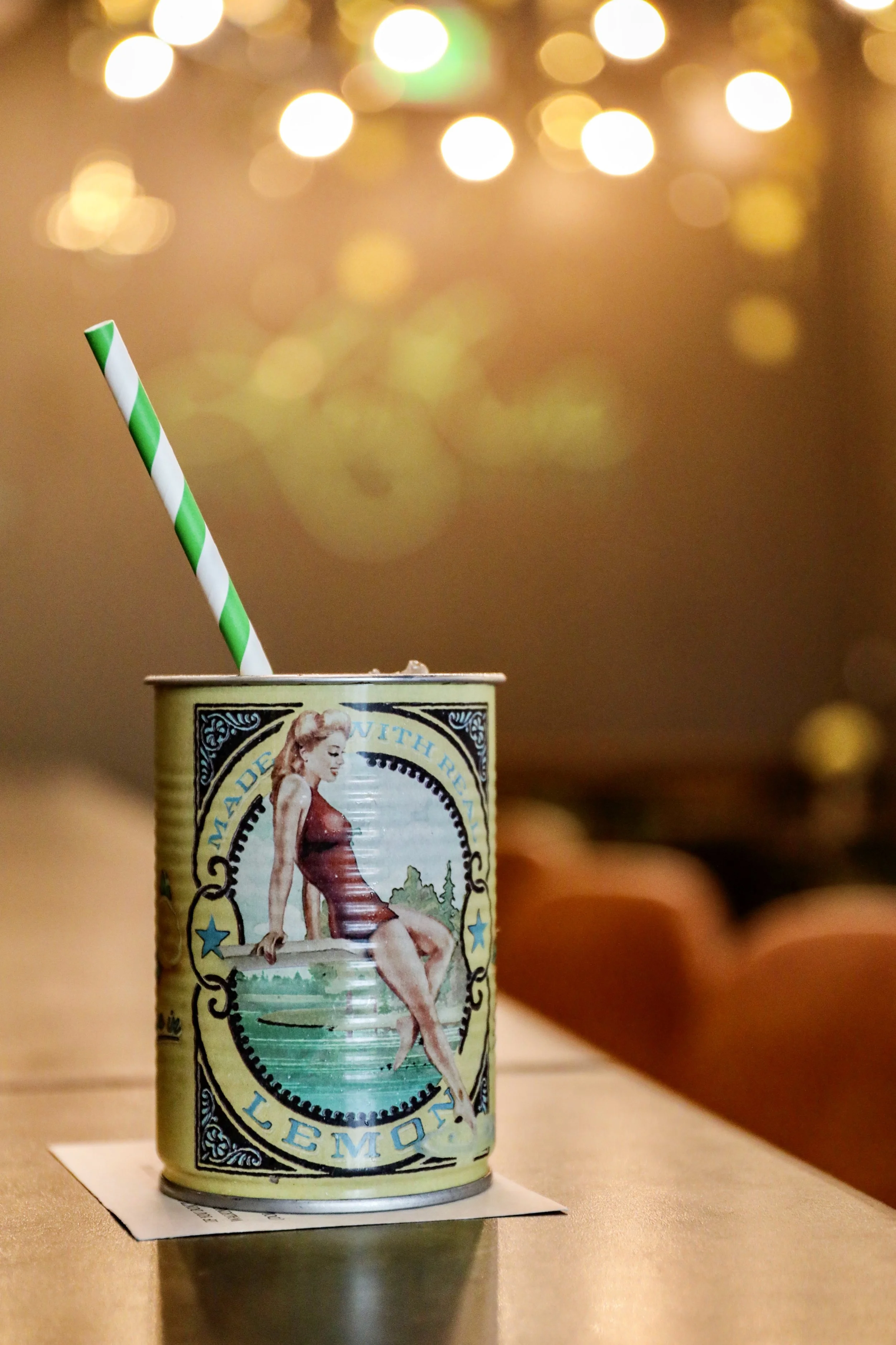 A can of Lemonade with a vintage pin-up girl illustration on the label. A green and white striped straw is inserted into the can, which is placed on a white paper on a wooden surface. Out-of-focus warm lights create a bokeh background.