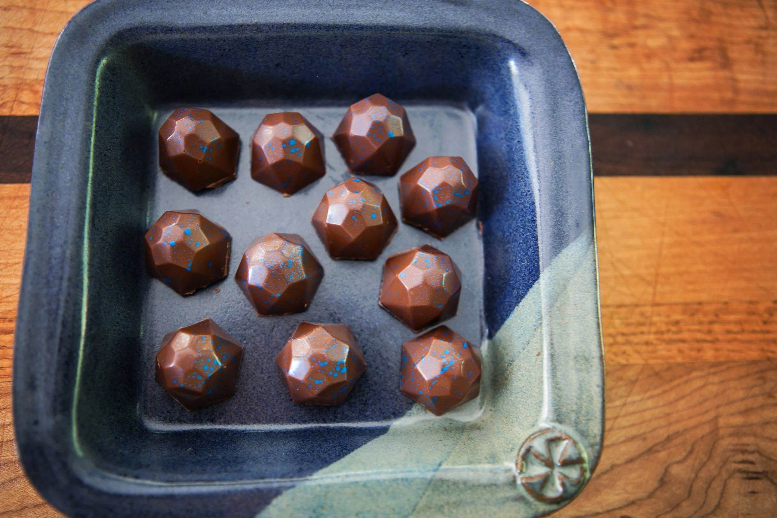 A small blue dish with eleven geometric-shaped chocolate truffles, each decorated with blue speckles, on a wooden surface.