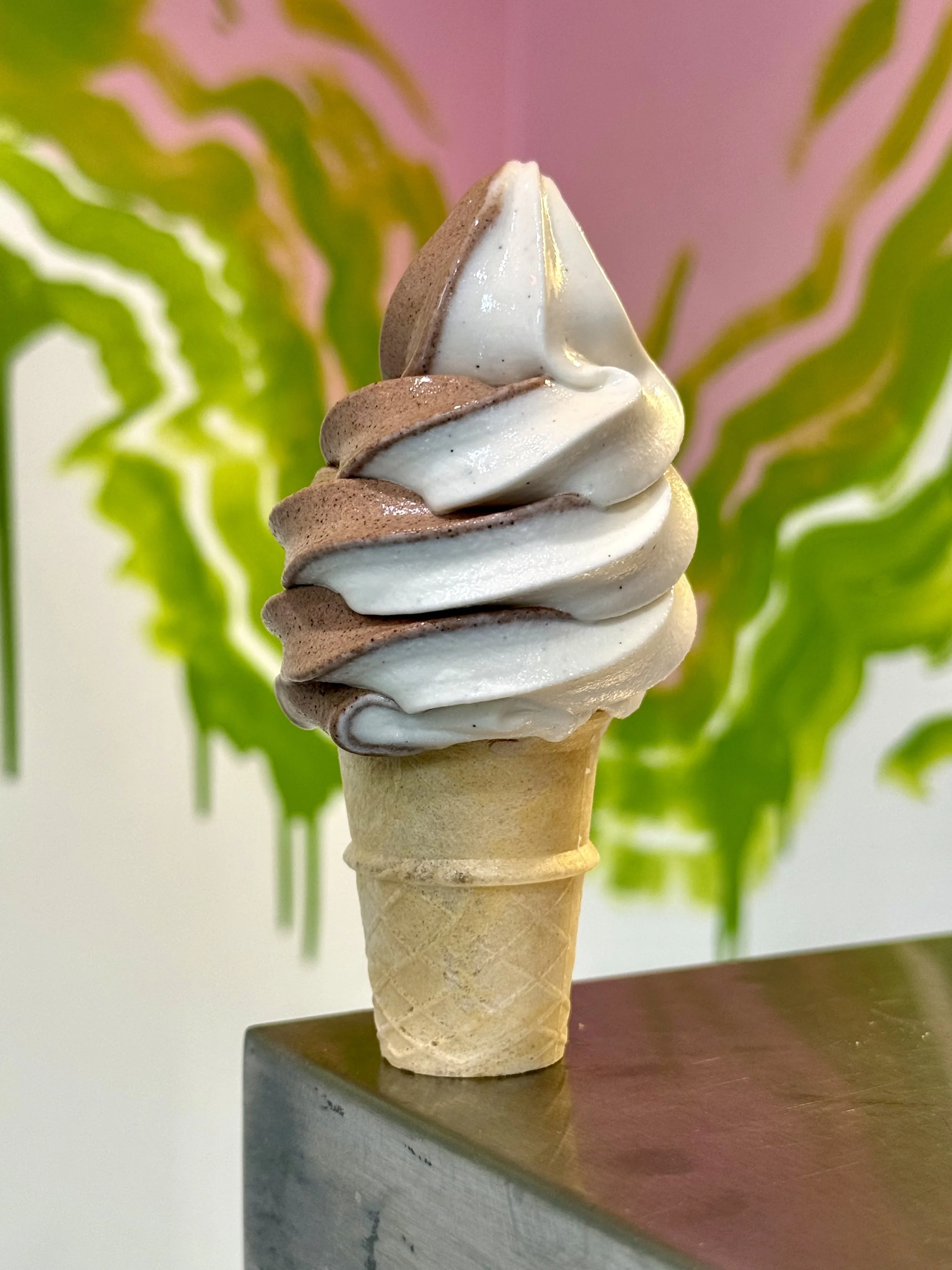 Close-up of a soft-serve ice cream cone with swirls of vanilla and chocolate, set against a colorful abstract background with green and pink hues.
