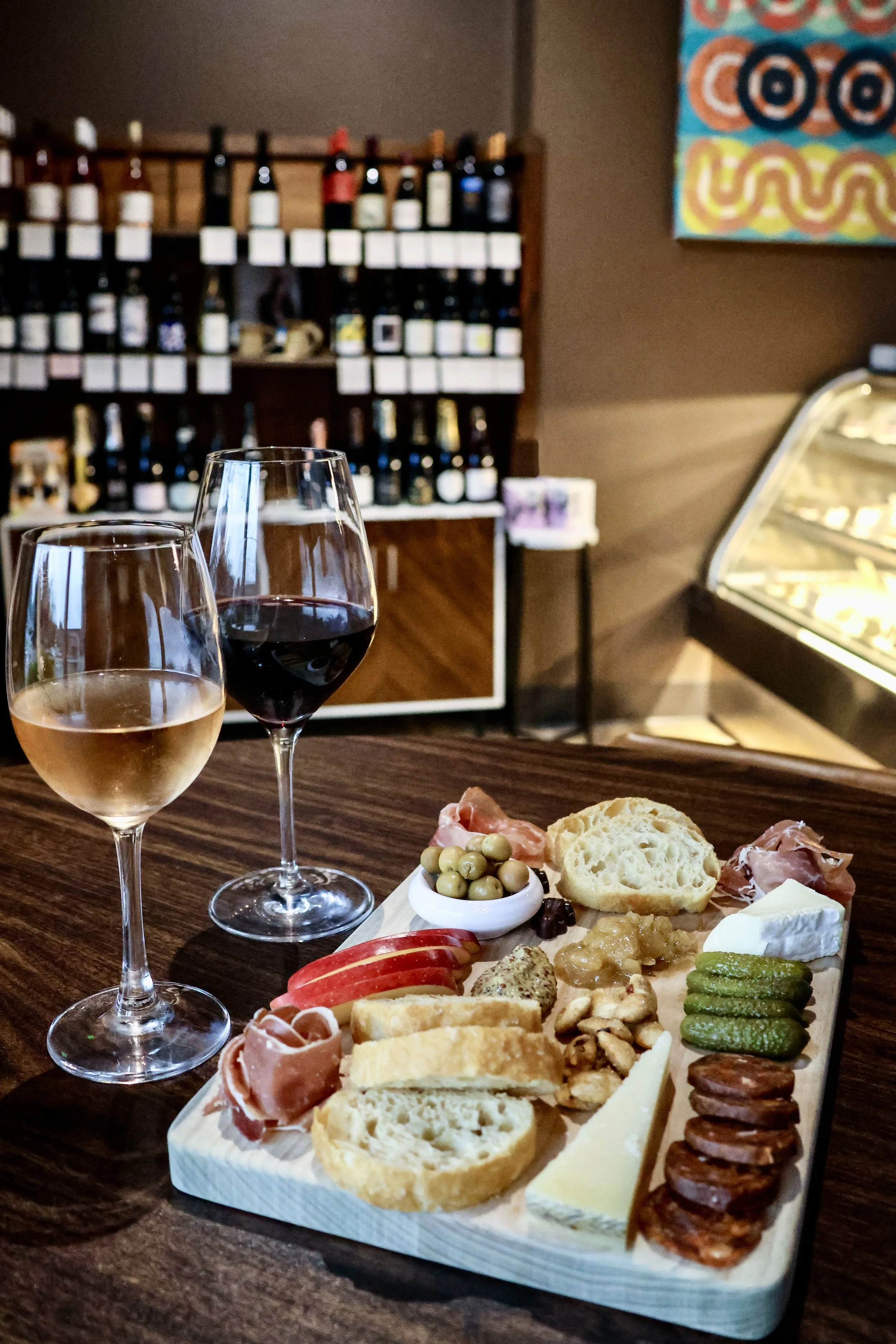 A charcuterie board with bread, cheese, meats, olives, and snacks, accompanied by two glasses of wine, set on a wooden table inside a cozy restaurant or cafe.