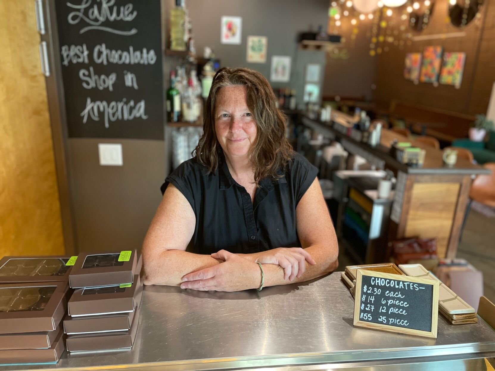 A smiling woman with curly hair sitting behind a counter in a chocolate shop. There are boxes of chocolates on the counter and a chalkboard sign listing prices for chocolates. The background features a menu board, bottles, and colorful wall decorations.