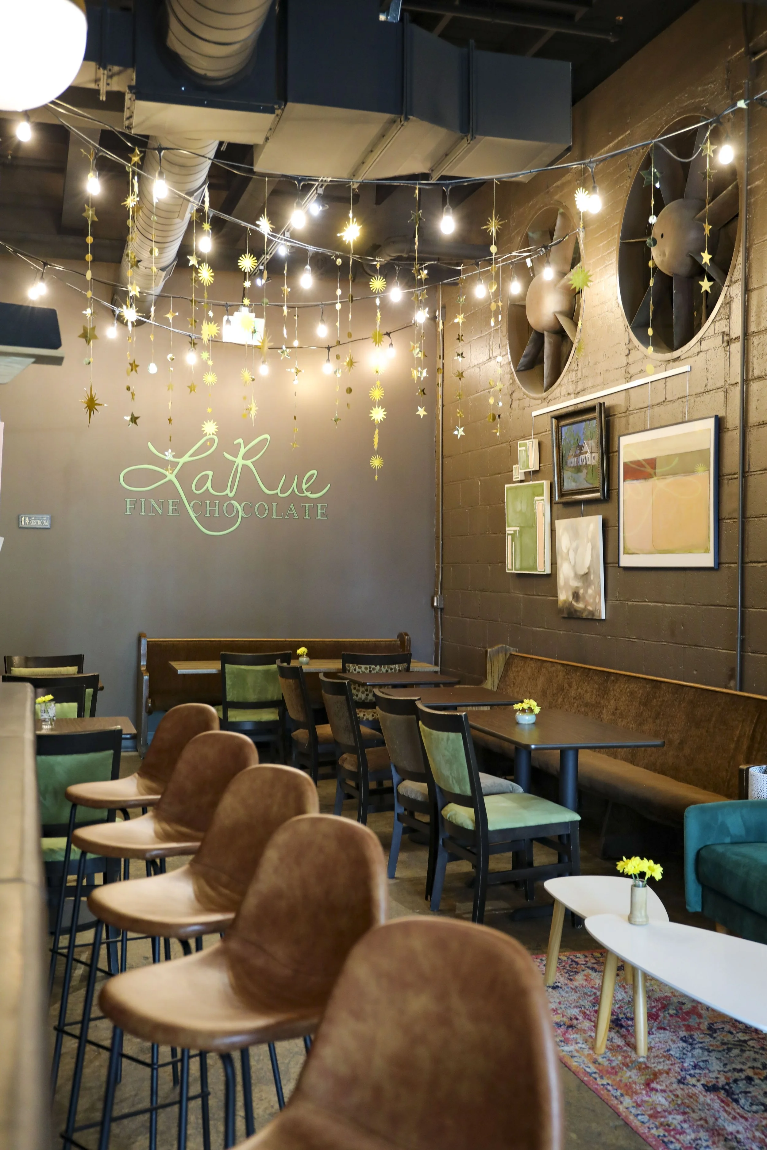 Interior of a coffee shop or cafe with wooden chairs, tables, and wall art including paintings and a large sign that reads 'La Rue Fine Chocolate'. Decorated with hanging star and sun-shaped ornaments and string lights.