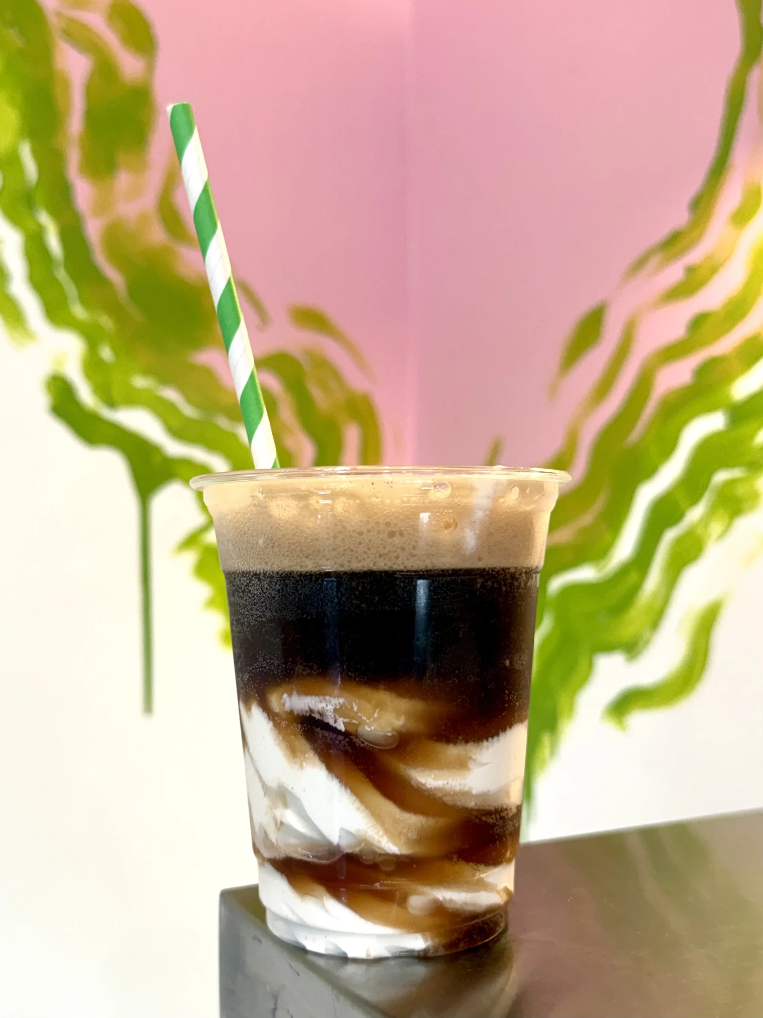 A tall iced coffee drink with layers of coffee, milk, and syrup in a clear plastic cup with a green and white striped straw, set on a dark surface with green leafy plants and a pink wall in the background.