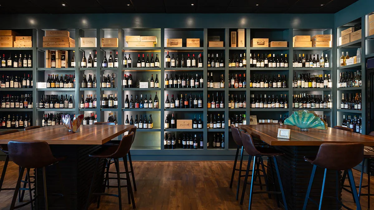 A wine shop with shelves filled with bottles of wine and wooden boxes, two tables surrounded by chairs, and decorative items on the tables.