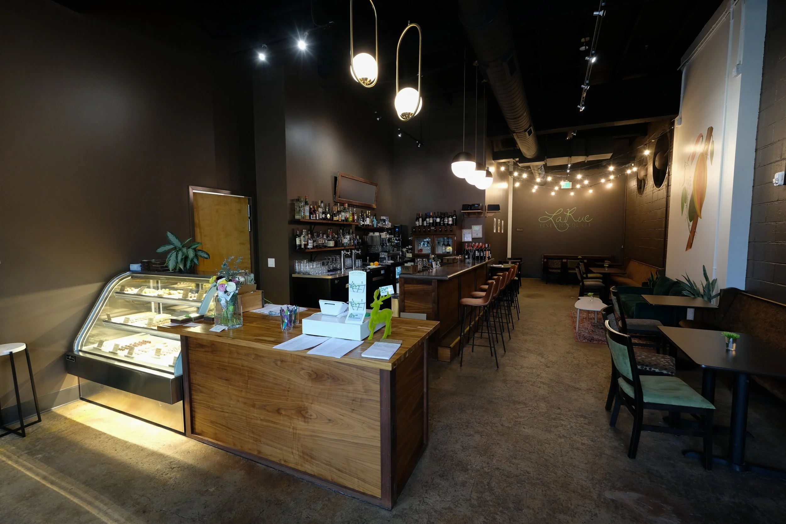 Interior of a modern cafe or bar with a wooden counter, bar stools, tables, and chairs, dim lighting, black walls, and decorative string lights, with a display case of desserts on the counter.