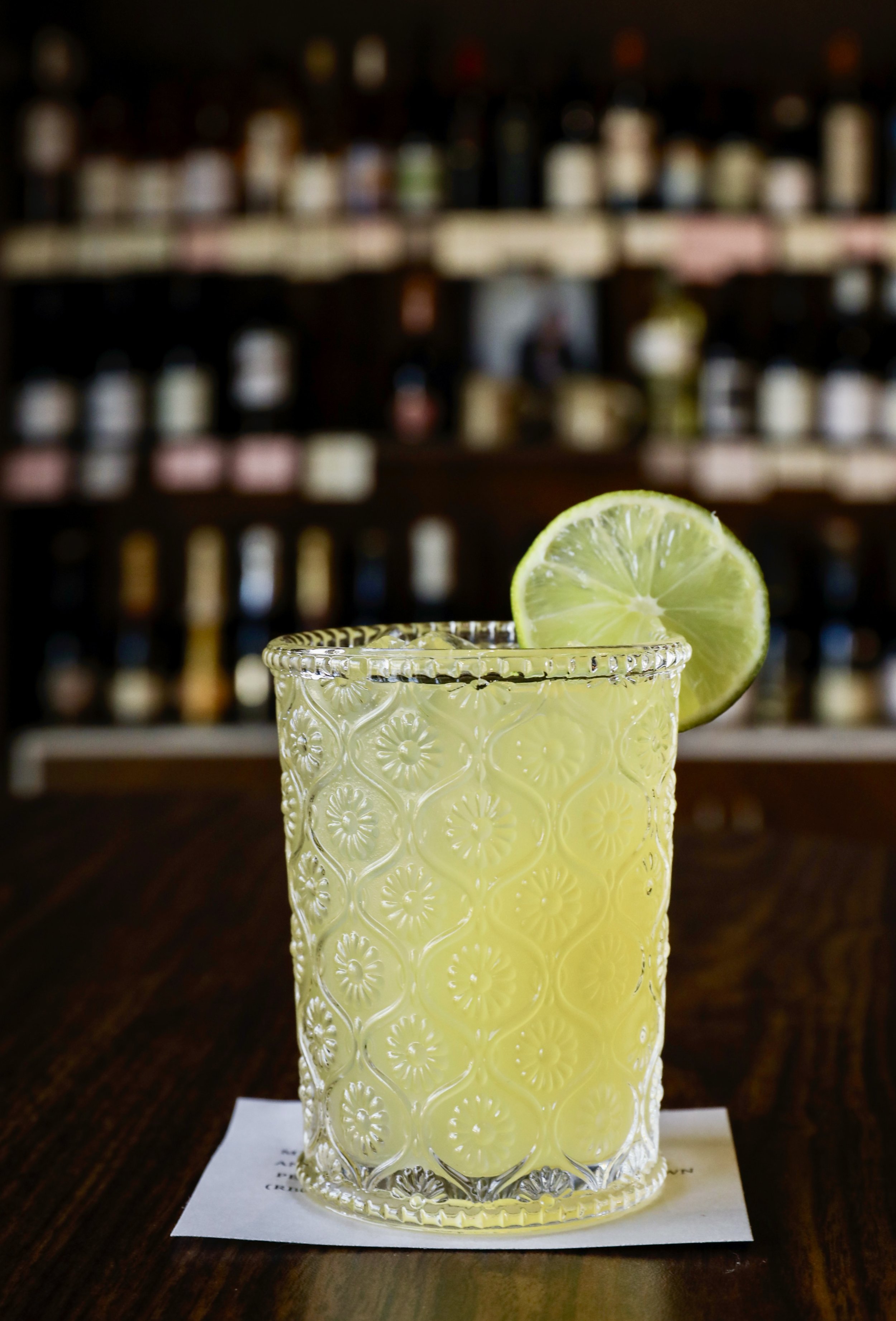A glass of yellow cocktail with a slice of lime on the rim, on a wooden table with a blurred background of bottles on shelves.