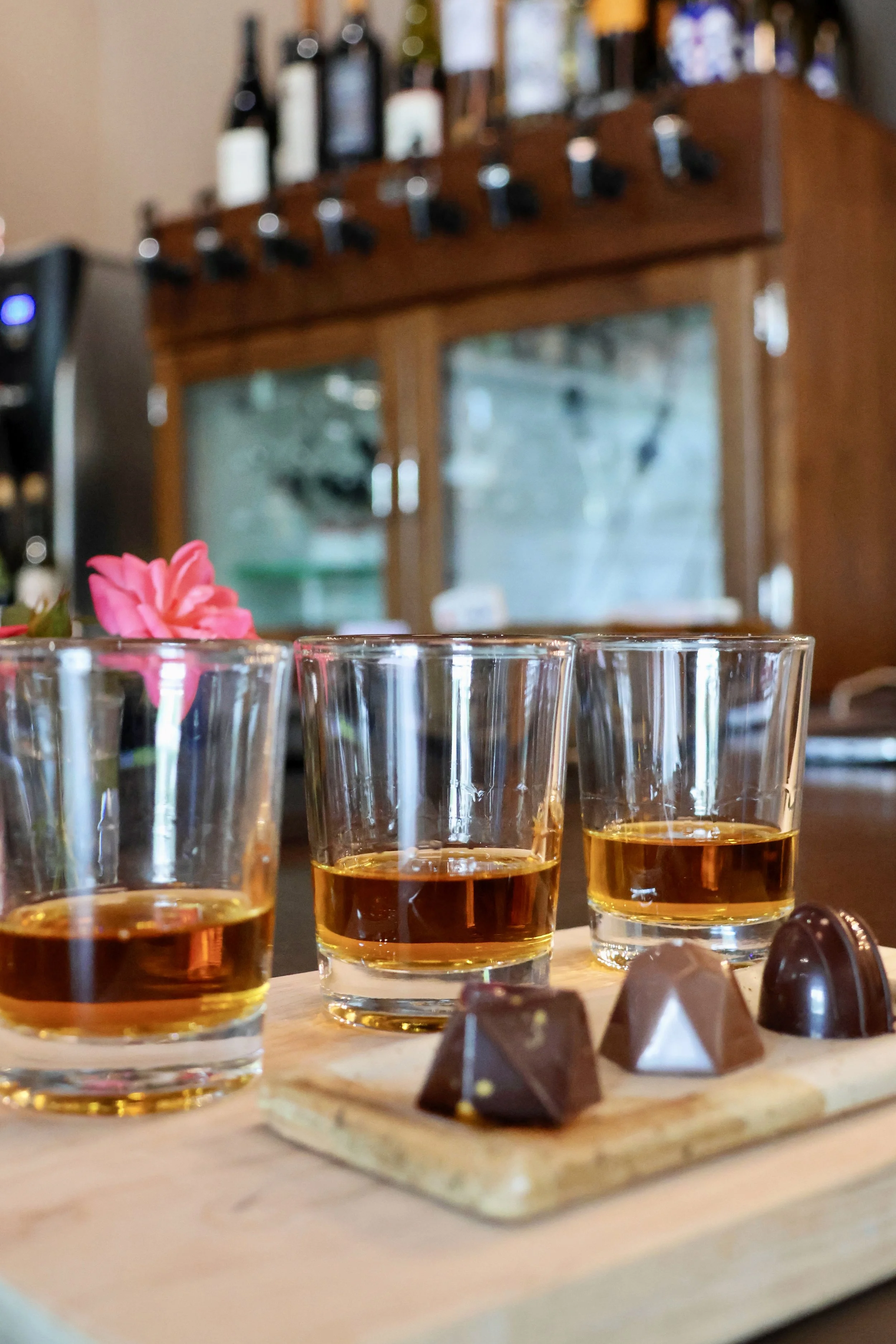 Three shot glasses filled with a golden-brown liquid, placed on a light-colored wooden board. To the left, a pink flower. To the right, four pieces of dark chocolate with nuts on top, on a small wooden board. In the background, a bar or shelf with liquor bottles, blurred.