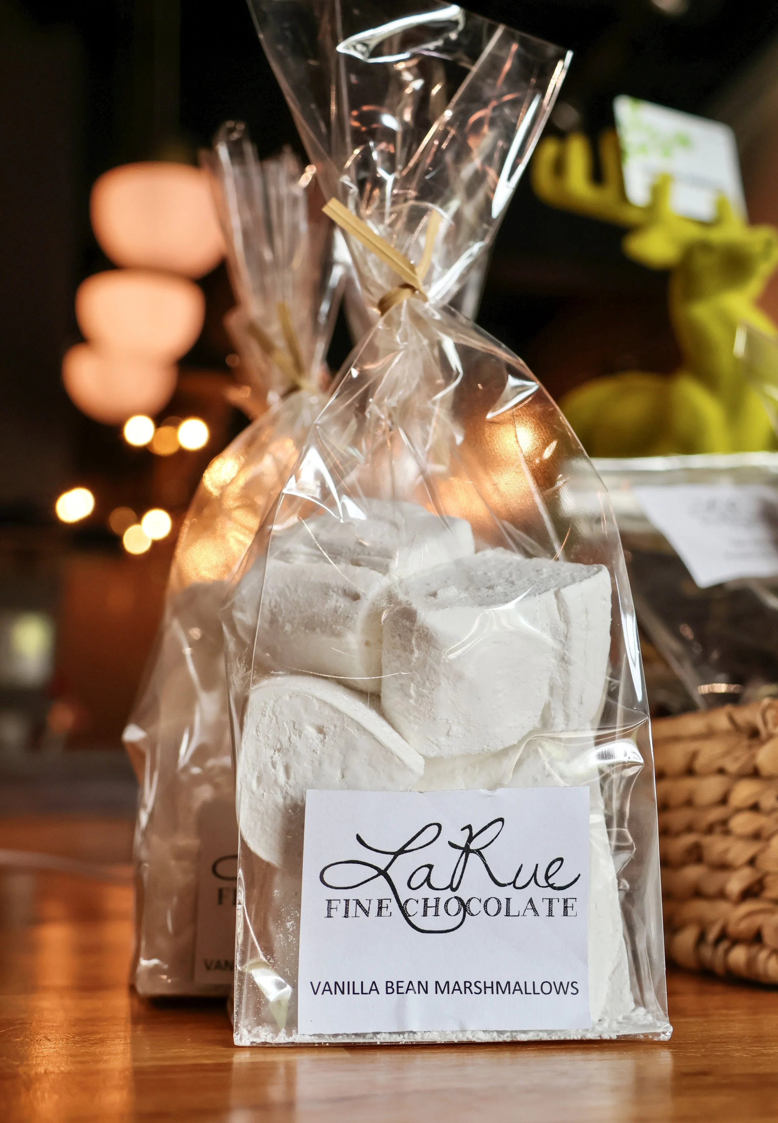 A clear plastic bag containing white vanilla bean marshmallows with a handwritten label that says 'LaRue' and 'VANILLA BEAN MARSHMALLOWS.' The bag is tied with a golden twist tie, and the background is blurred with warm lighting.