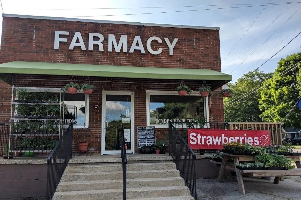 Brick building with a large sign that reads 'FARMACY.' Potted plants hang under a green awning, and a red banner advertises 'Strawberries' near outdoor display tables with plants and flowers.