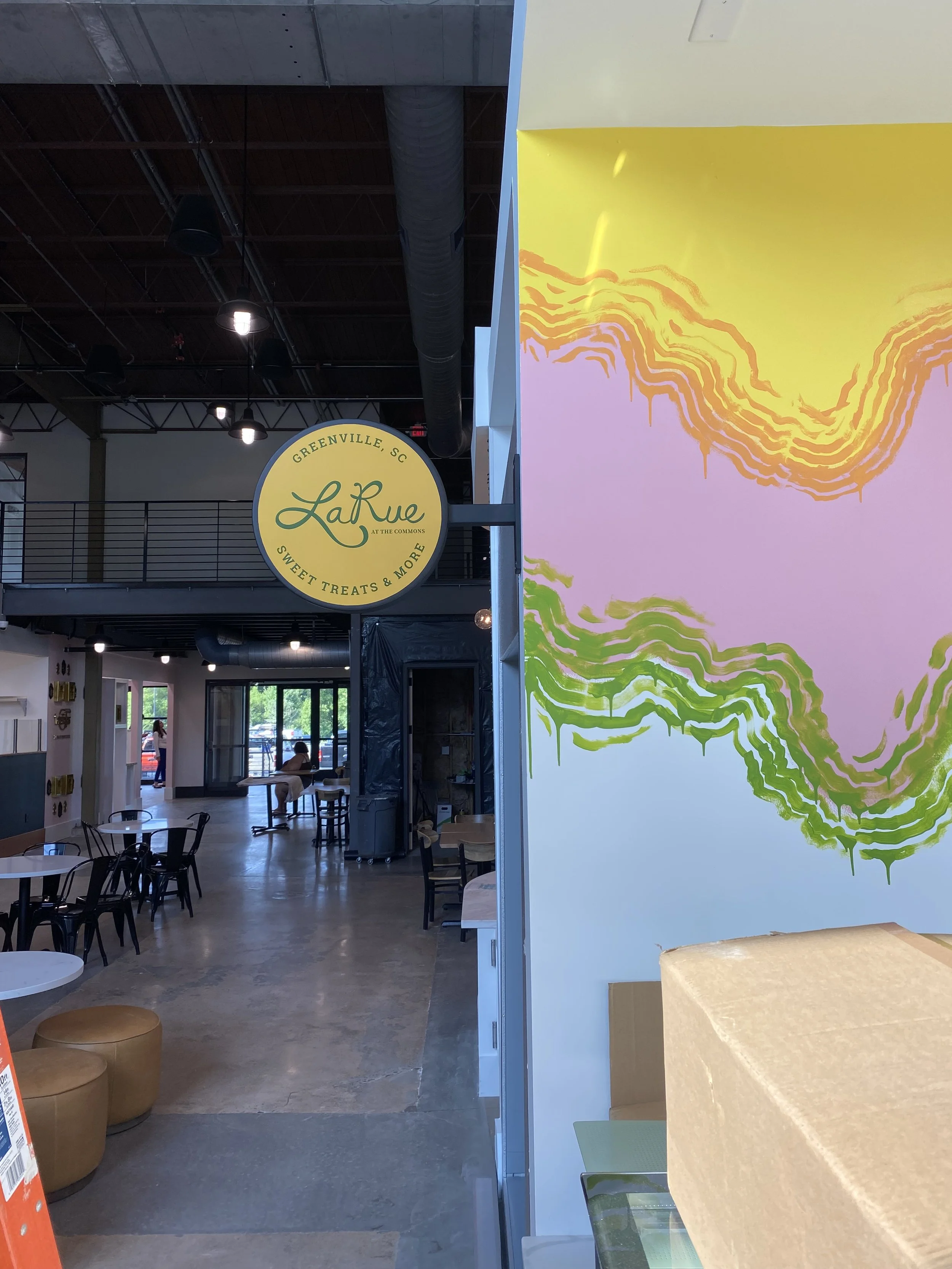 Interior of a modern cafe or restaurant with tables and chairs, a colorful abstract wall art, and a circular yellow sign reading 'LaRue' at The Commons in Greenville, SC, with visible ceiling ductwork and hanging lights.