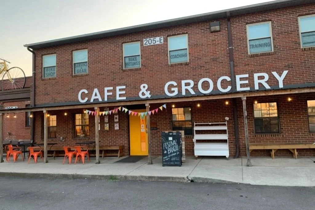 Exterior of a brick building with a large sign that reads 'Café & Grocery', outdoor seating with orange chairs, a yellow door, festive bunting, and a chalkboard sign advertising the Brazil Festival.