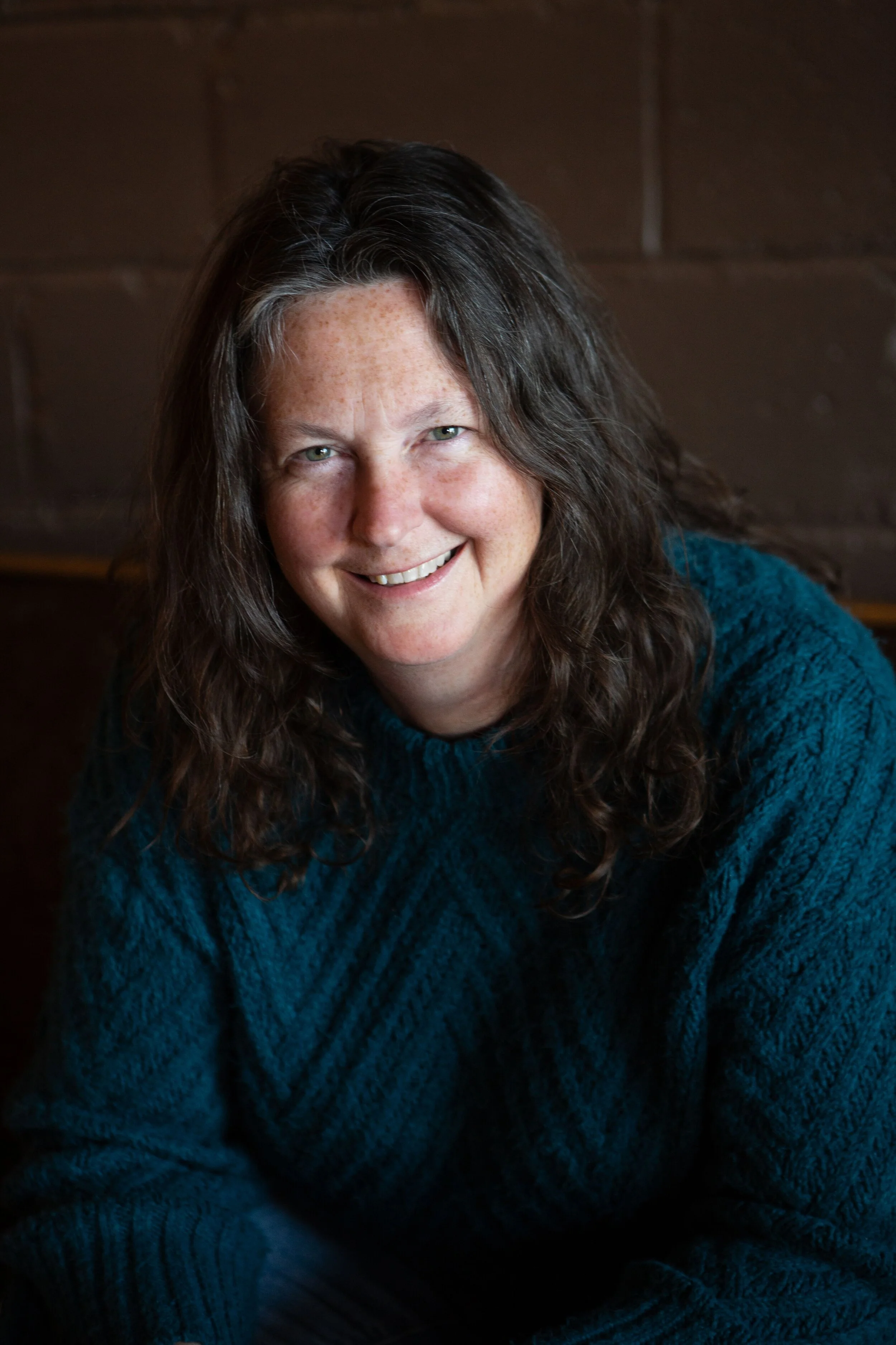 Close-up of a woman with long wavy dark hair, light skin with freckles, smiling, wearing a teal sweater, sitting in front of a brown background.