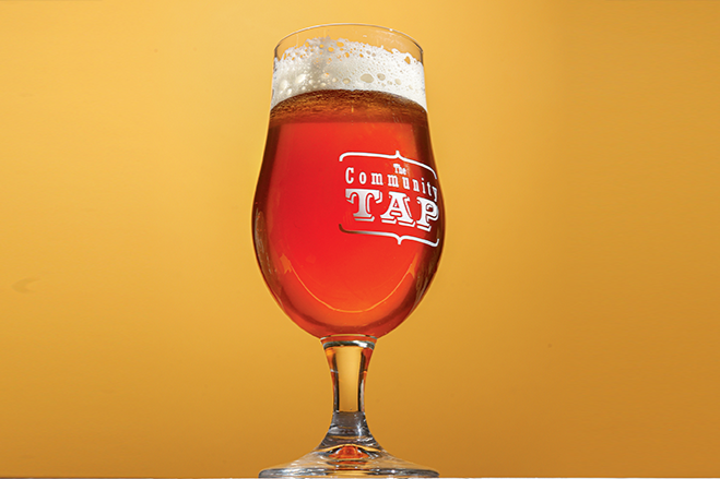 A beer glass filled with amber-colored beer with a foamy head, placed against a yellow background, with The Community Tap logo on the glass.