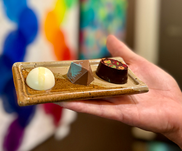 Hand holding a small ceramic plate with three pieces of assorted chocolates, against a colorful, blurred background.