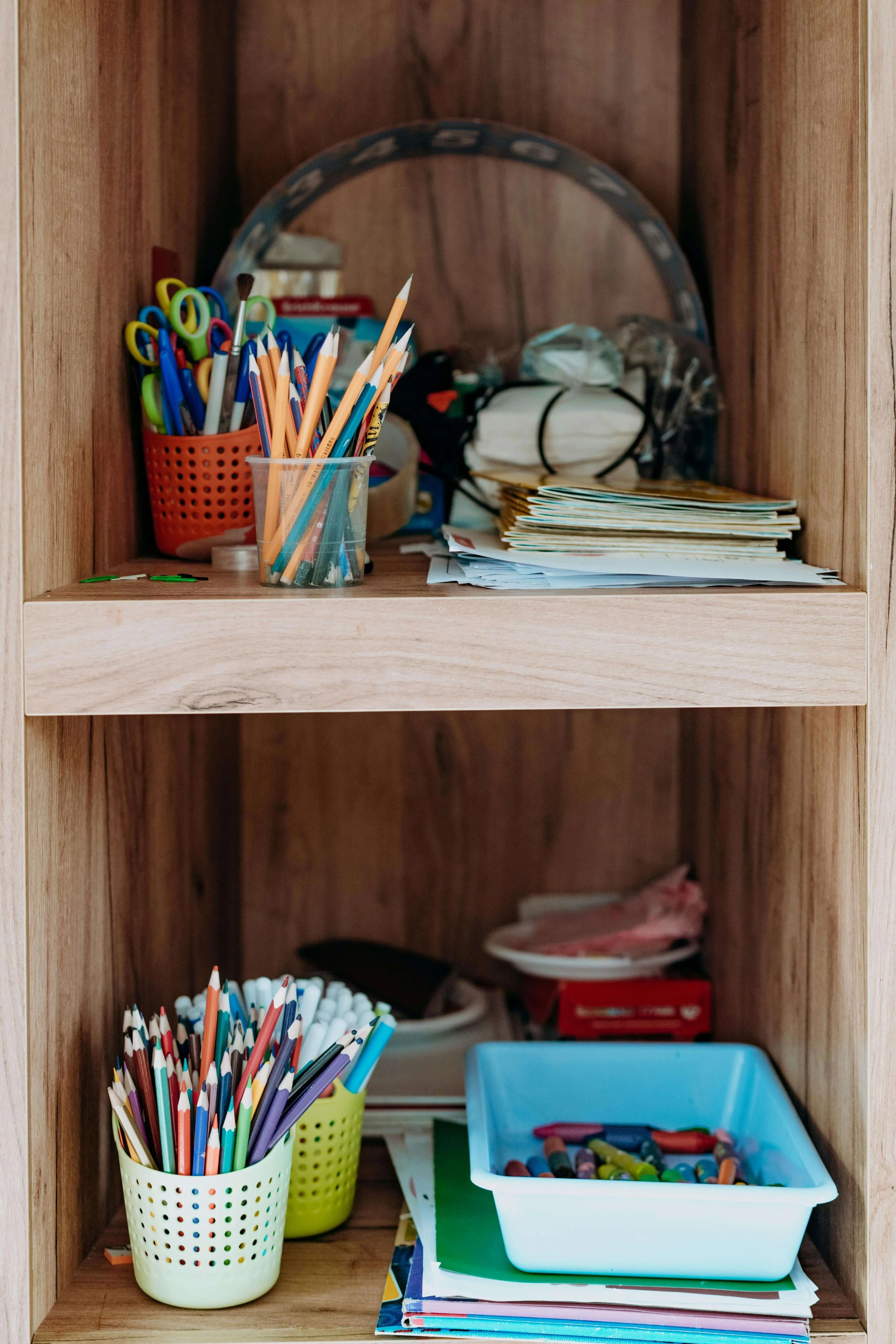 A wooden shelf with school supplies including colored pencils, markers, and scissors organized in containers, along with notebooks and crayons.