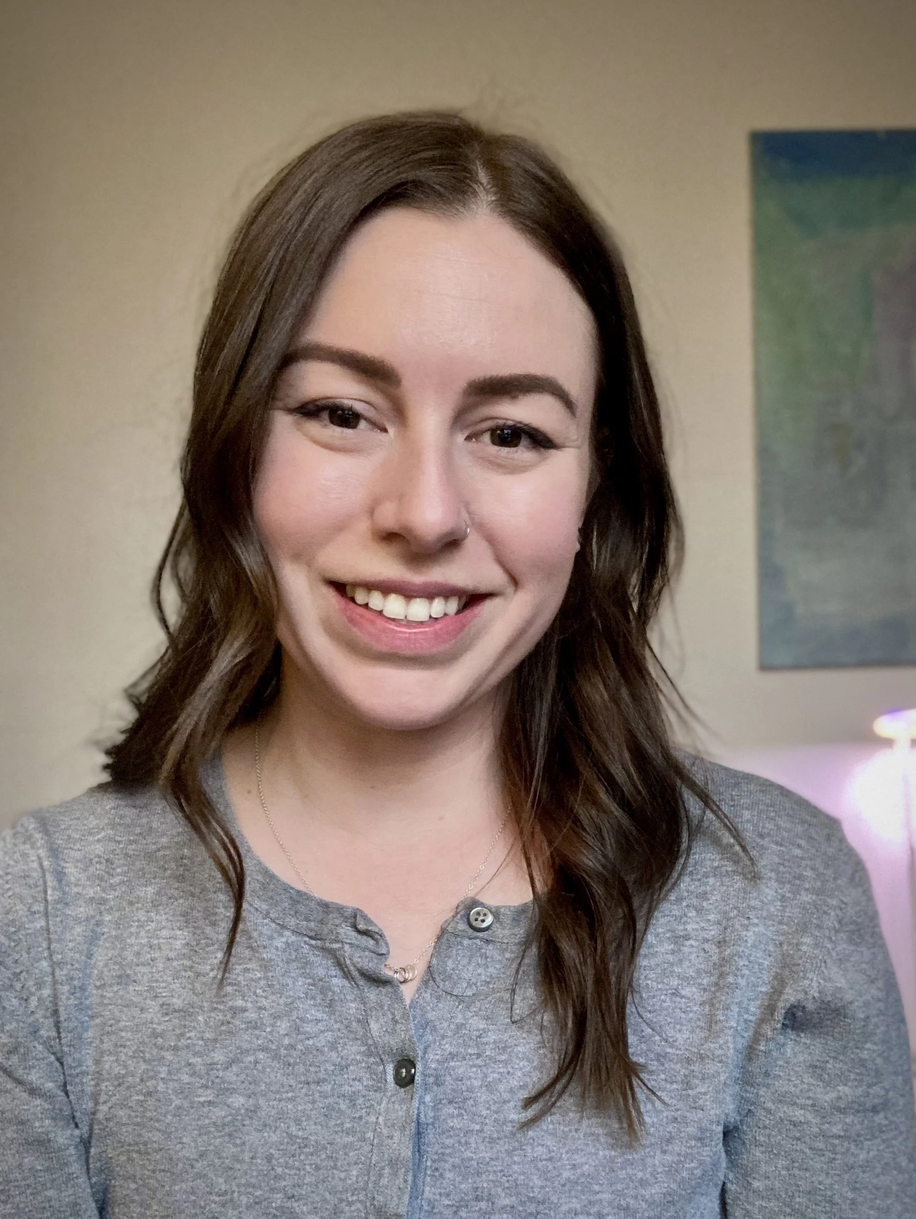 Young woman with long brown hair smiling, wearing a gray button-up shirt, standing indoors near a beige wall and a colorful painting.