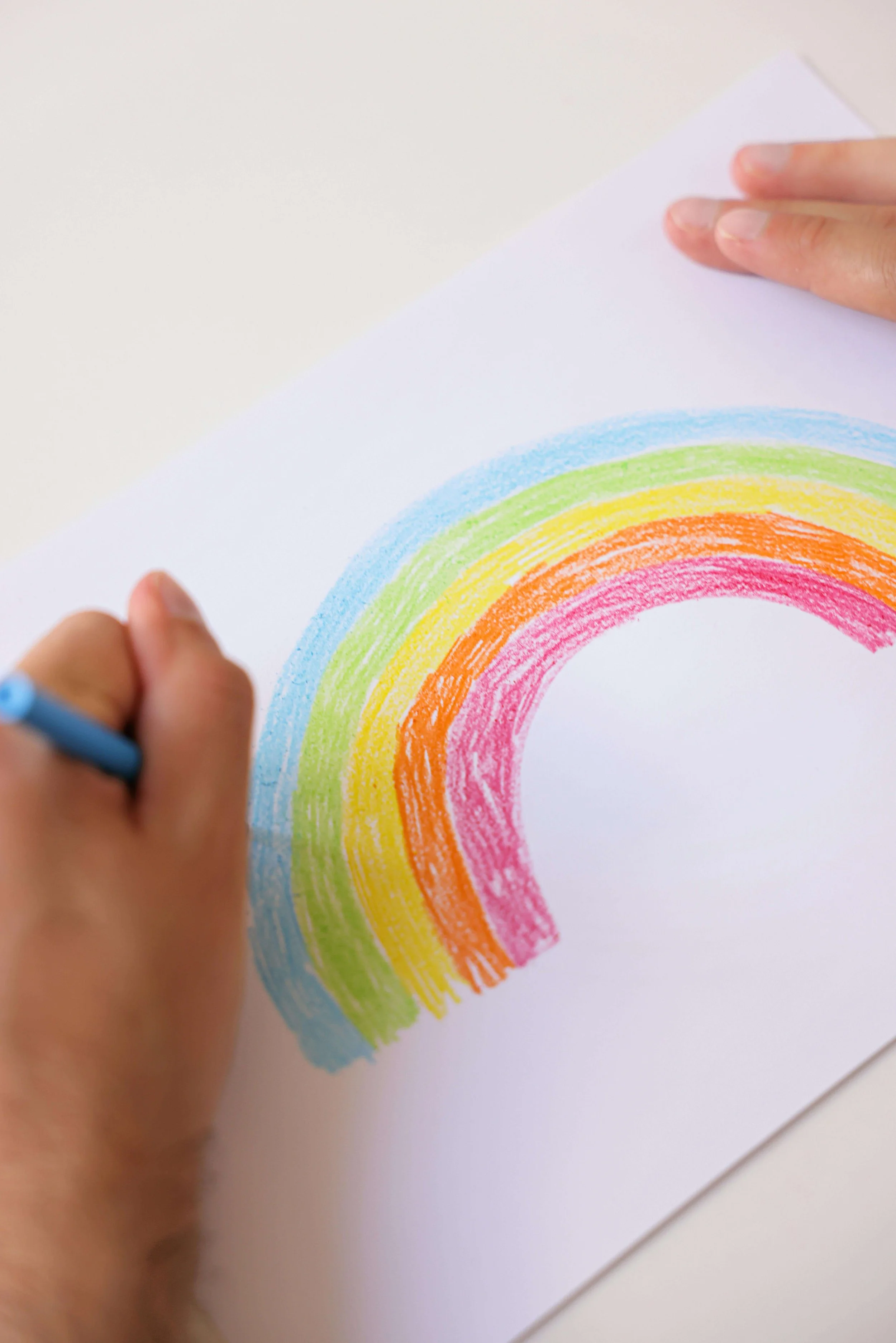 Child coloring a rainbow with crayons on white paper.