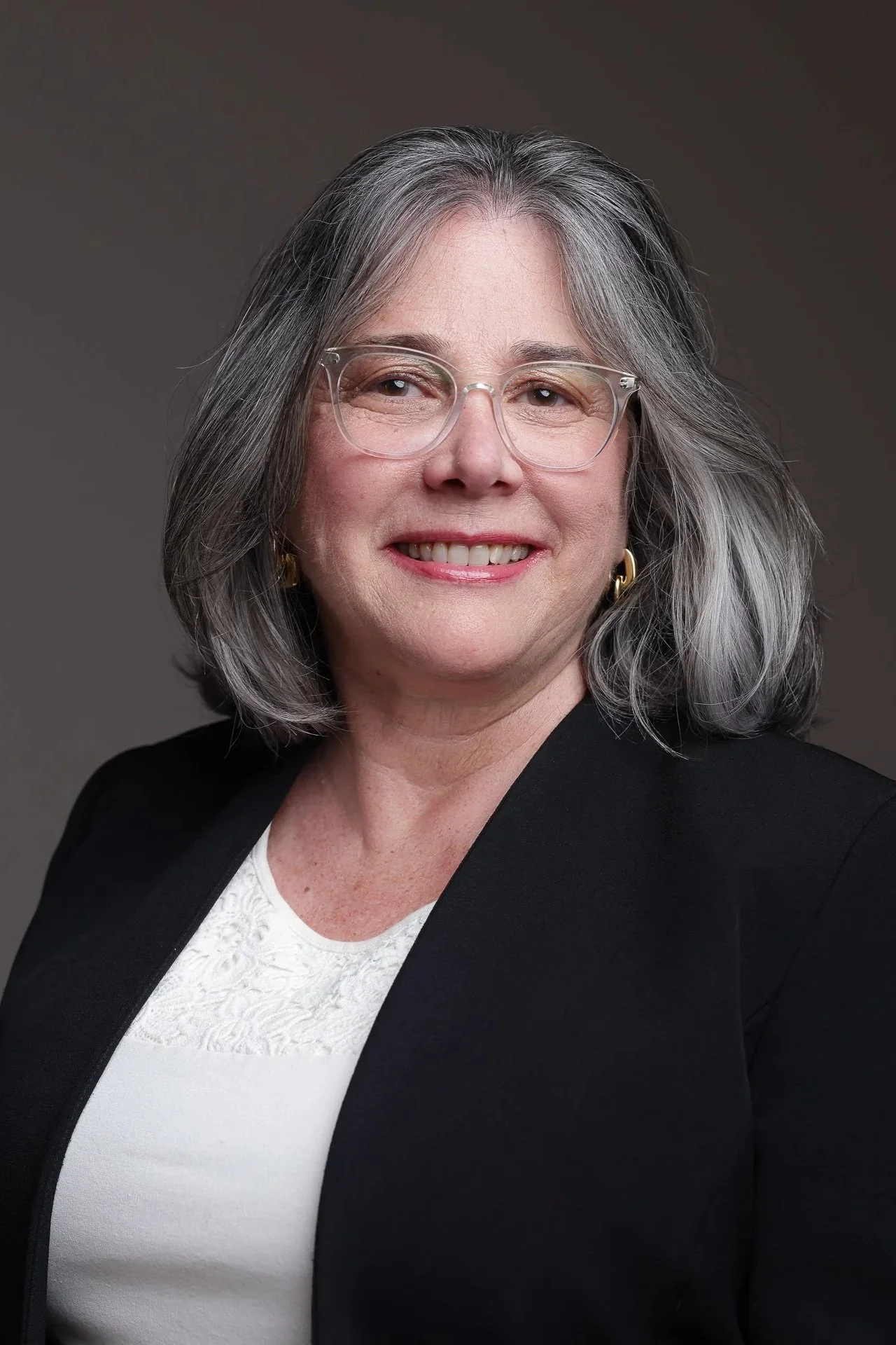A portrait of Ann McCulloch, Executive Producer of 'Shall Men Vote?' film and great-granddaughter of Catharine Waugh McCulloch. She is wearing clear glasses, gold earrings, a white top, and a black blazer, smiling against a dark gray background.