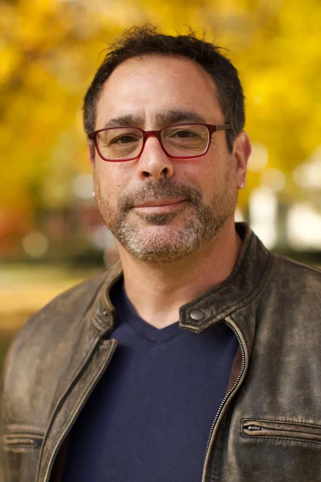A portrait of Dean Bushala, Producer of 'Shall Men Vote?' film. He is wearing glasses, has dark hair and a beard, wearing a brown leather jacket and a navy shirt, standing outdoors with a background of fall foliage.