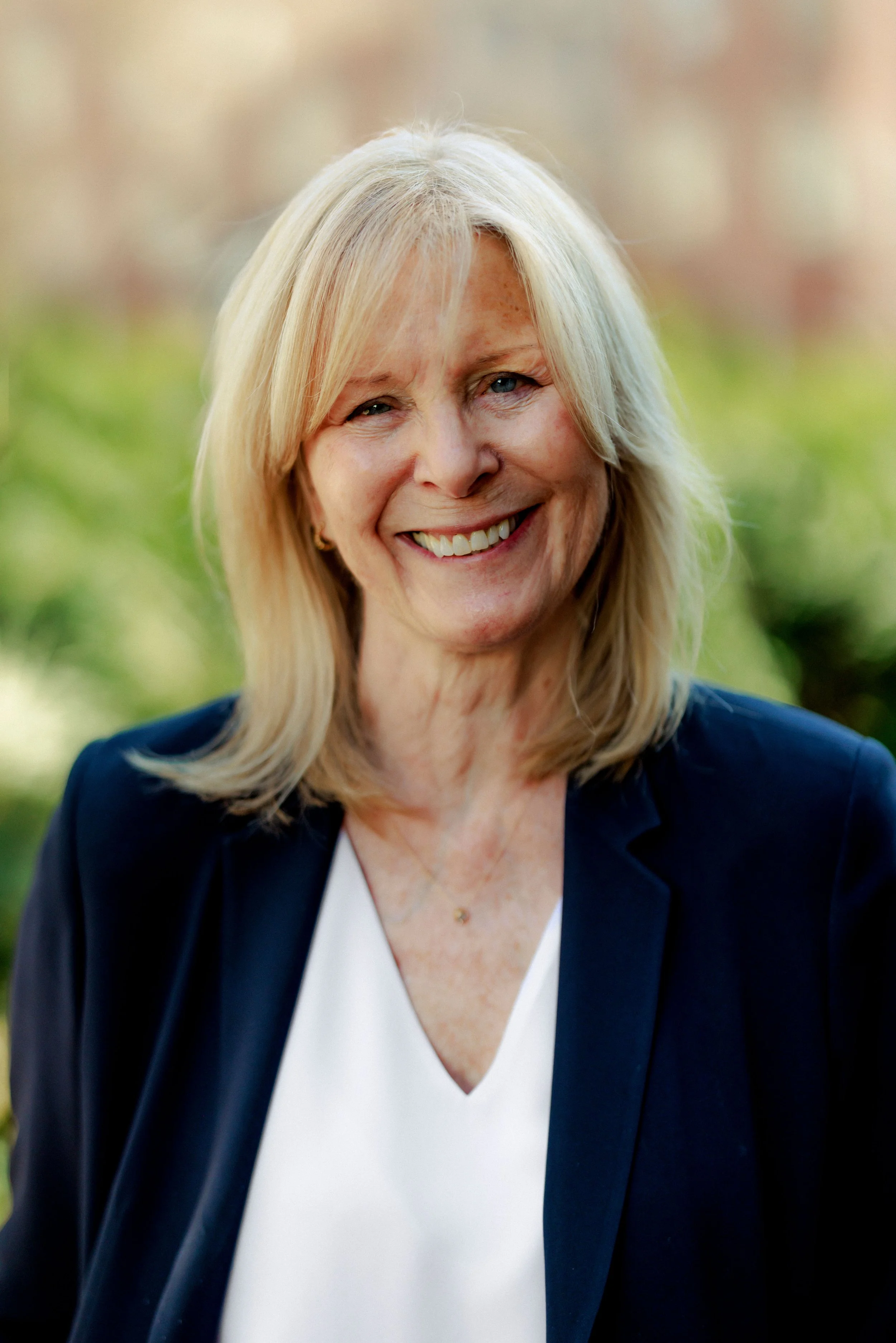 A portrait of Susan Hope Engel, Director and Editor of 'Shall Men Vote?' film. She has blonde hair, wearing a navy blazer and a white top, and standing outdoors with a blurred natural background.