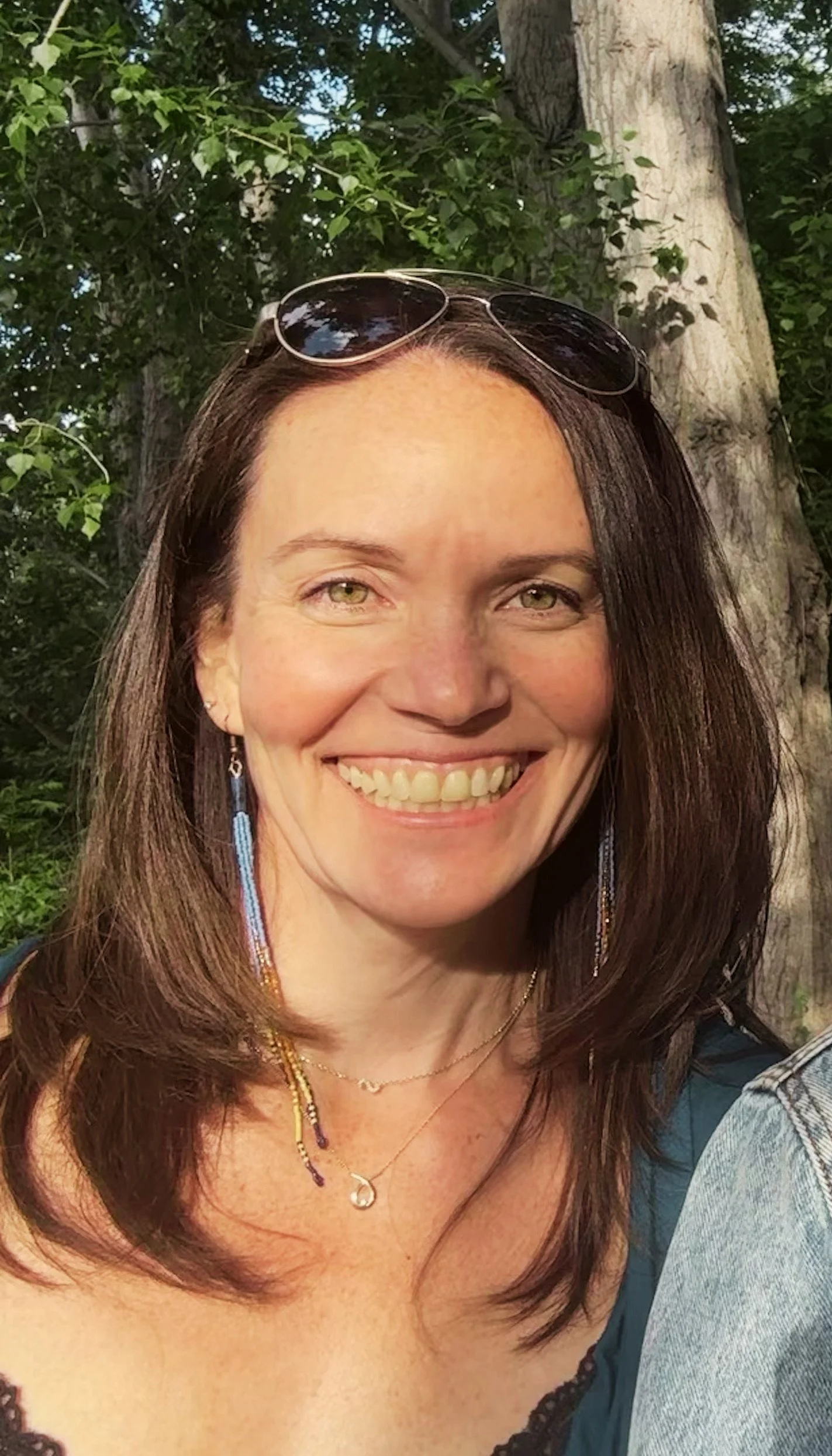 A smiling woman with long brown hair wearing sunglasses on her head, jewelry, and a blue top, outdoors with trees in the background.