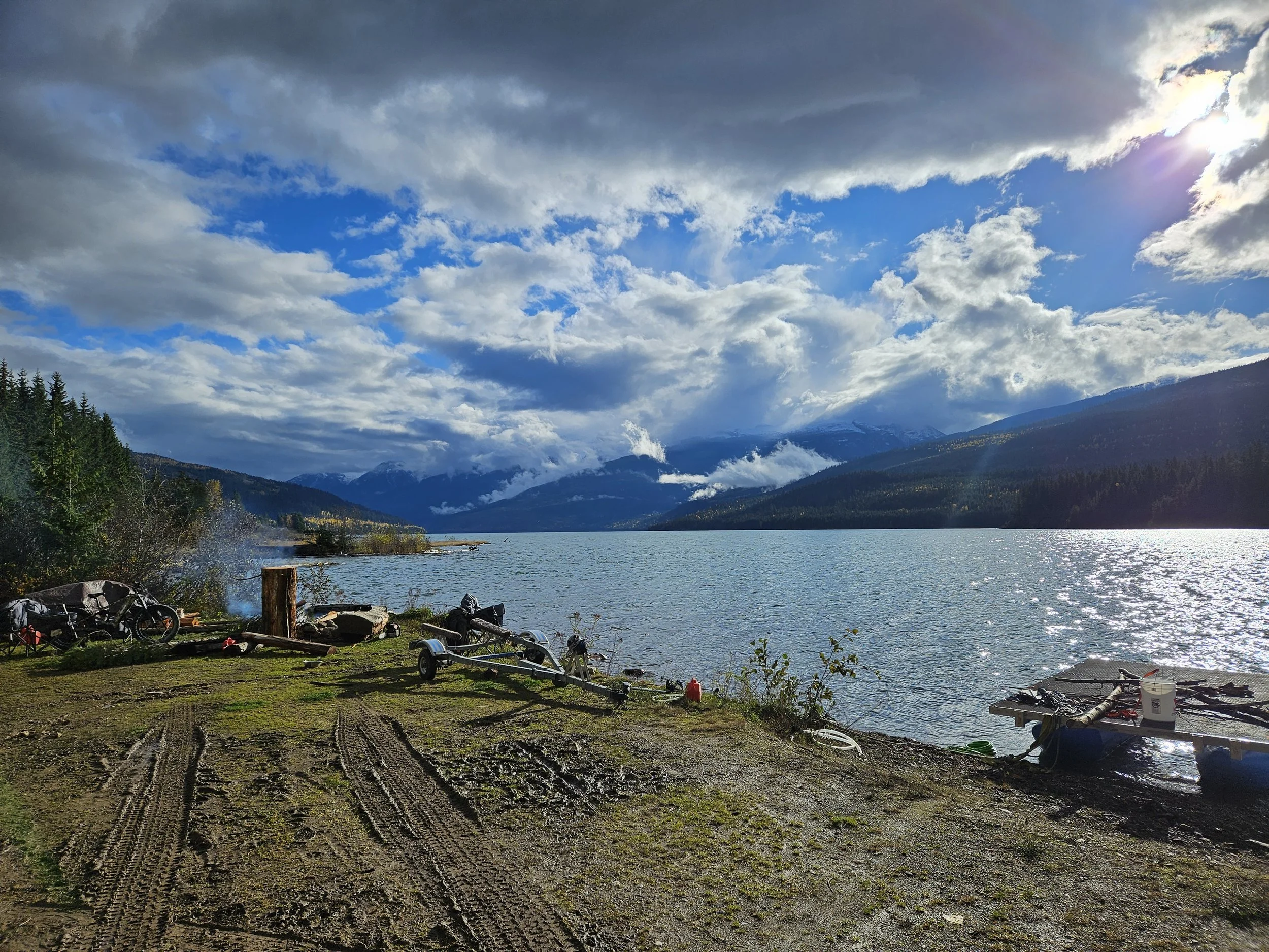 A lakeside view with mountains in the background, partly cloudy sky with sunlight peeking through, and a grassy foreground with parked boats and equipment.