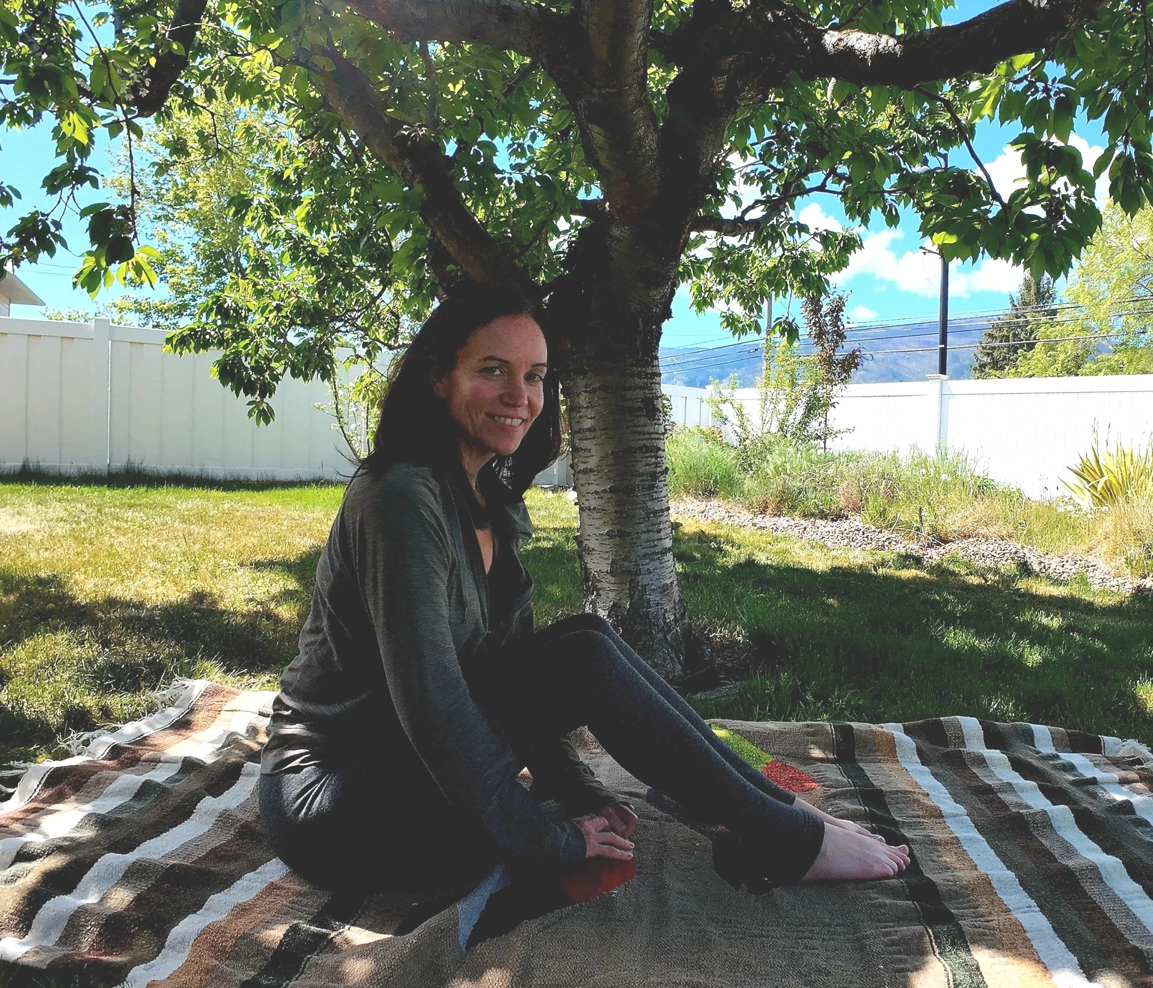 A woman sitting under a tree on a blanket in a backyard, smiling at the camera, with a white fence, blue sky, and mountains in the background.