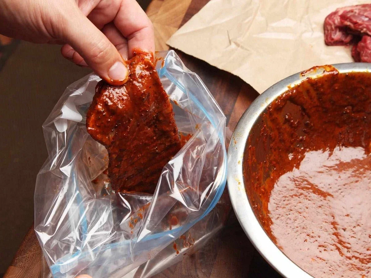 Person holding a seasoned piece of raw meat, likely pork, in a clear plastic bag. A large mixing bowl with red marinade or sauce is nearby on a wooden surface.
