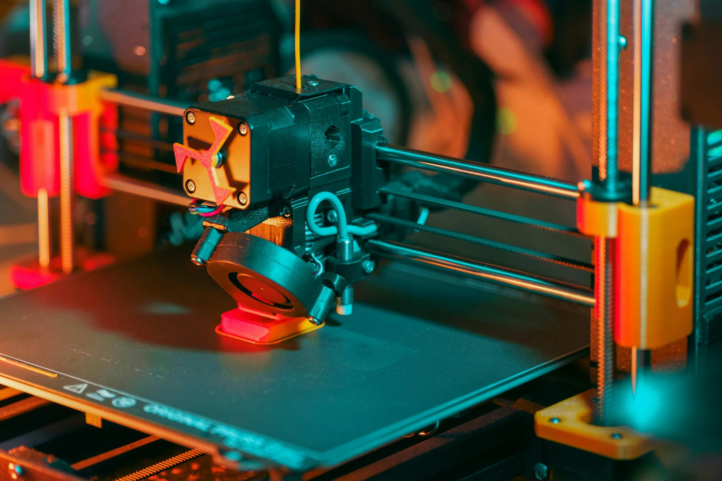 Close-up view of a 3D printer's extruder and print bed, with various colored parts and components in focus.
