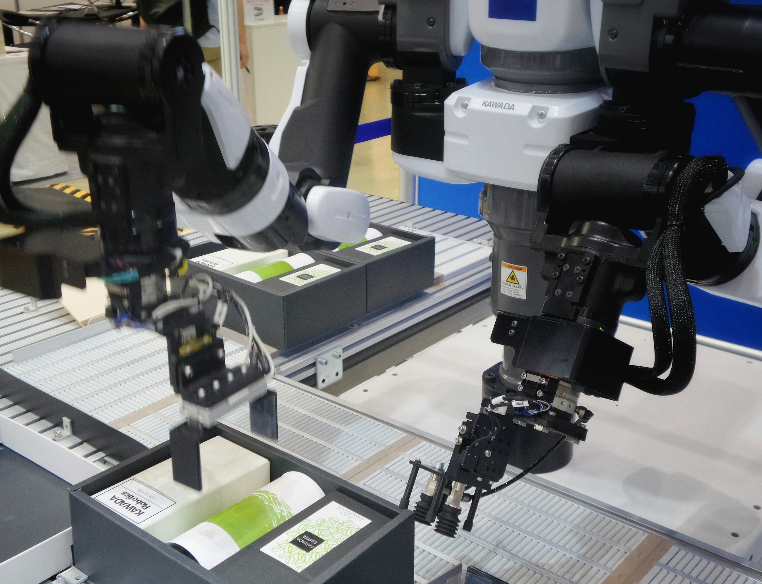 Robotic arm working on a project at an exhibition or workshop, with various displayed items and informational sheets below.