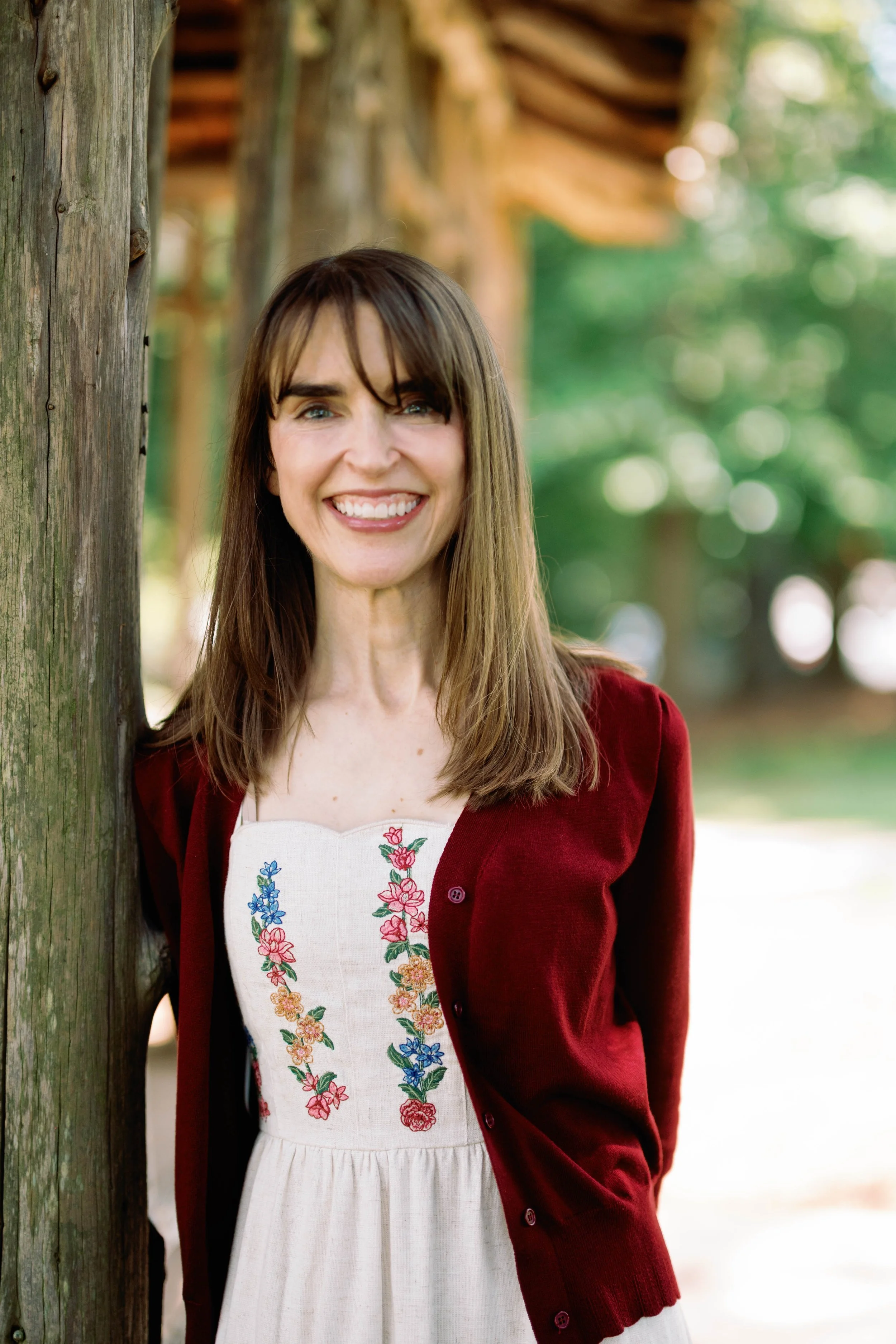 A woman with long brown hair, blue eyes, and a bright smile standing outdoors next to a wooden post, with blurred green trees and sunlight in the background. She wears a white dress with embroidered floral patterns and a red cardigan.