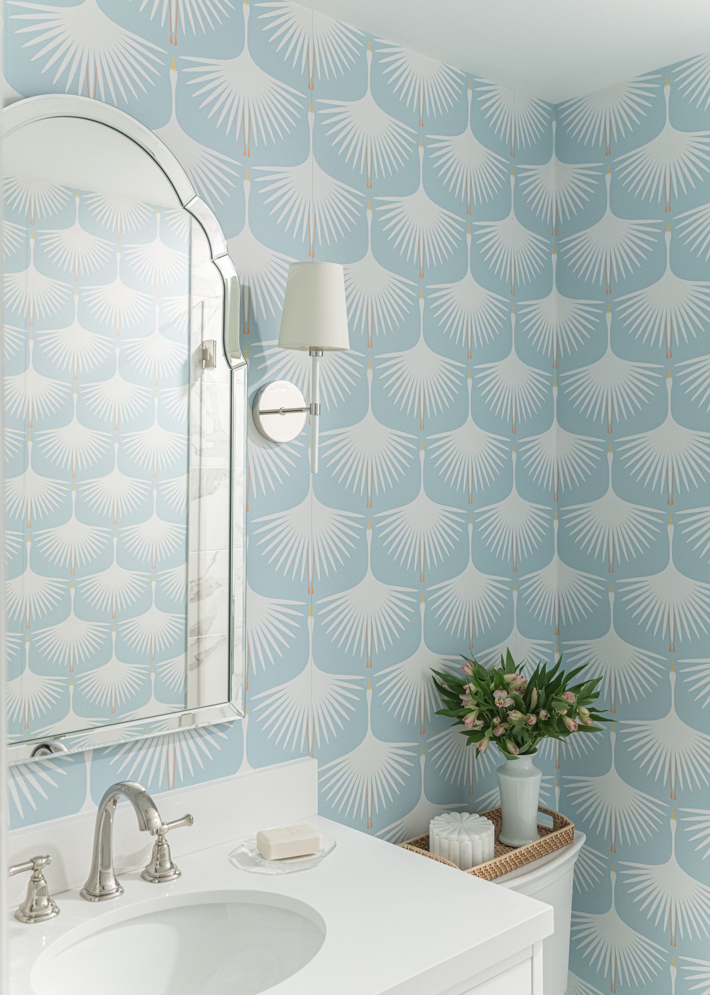 Bathroom with a mirror, white sink, silver faucet, soap bar, white tissue, blue patterned tile wall, white lamp, vase with pink flowers, small round basket, and a wicker storage unit.