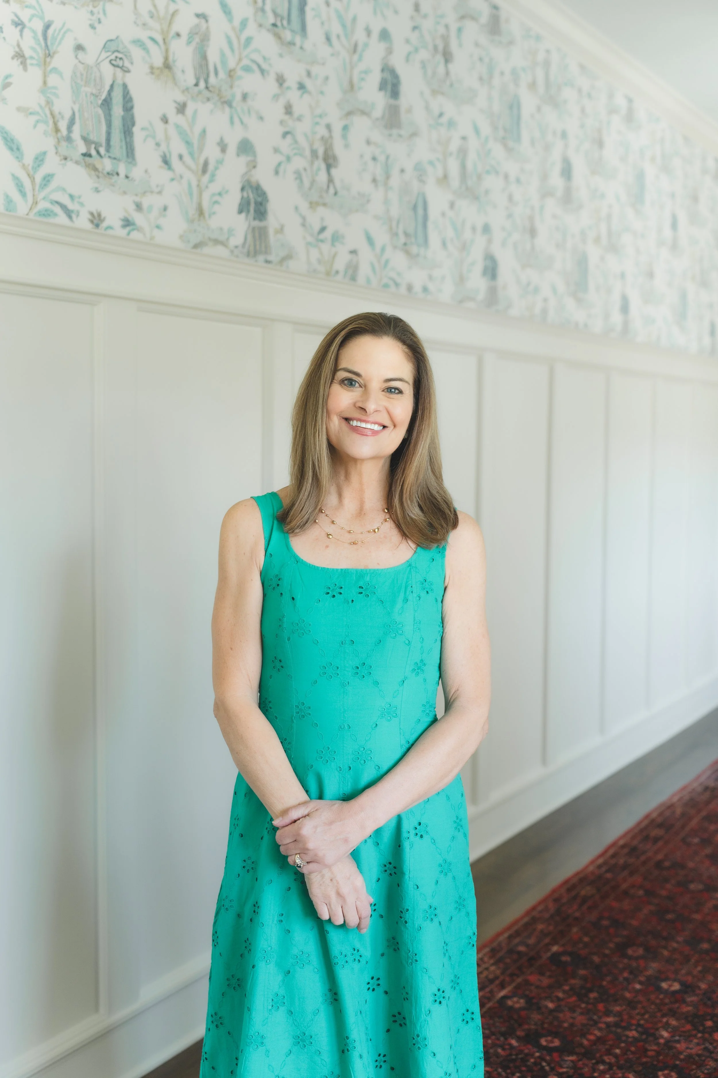 A woman with shoulder-length brown hair smiling, wearing a sleeveless turquoise dress with eyelet details, standing in front of a white paneled wall with patterned wallpaper. Her hands are clasped in front of her.