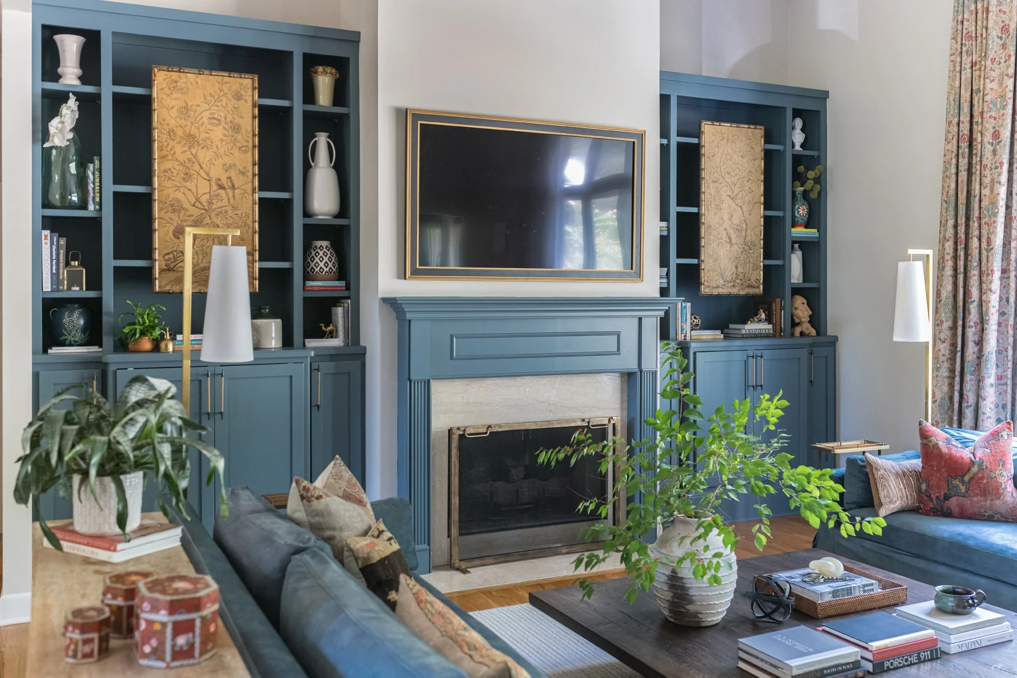 Living room with blue built-in bookshelves on either side of a fireplace, a large flat-screen TV, a gray couch with patterned pillows, a wooden coffee table with books, a large potted plant, and decorative items including vases and framed art.
