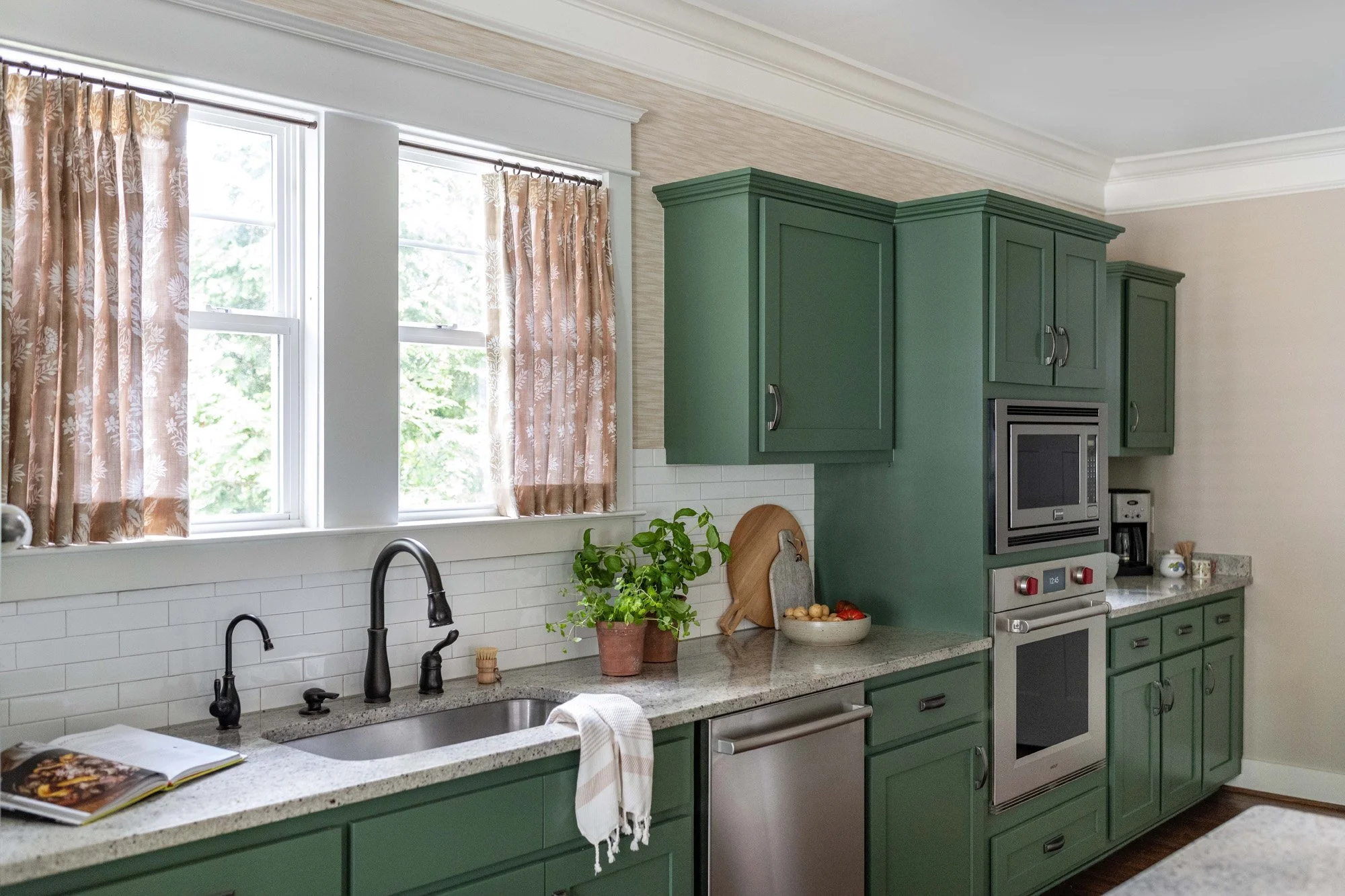 Kitchen with green cabinets, stainless steel oven, white subway tile backsplash, black faucet, and beige curtains with floral pattern at the windows.