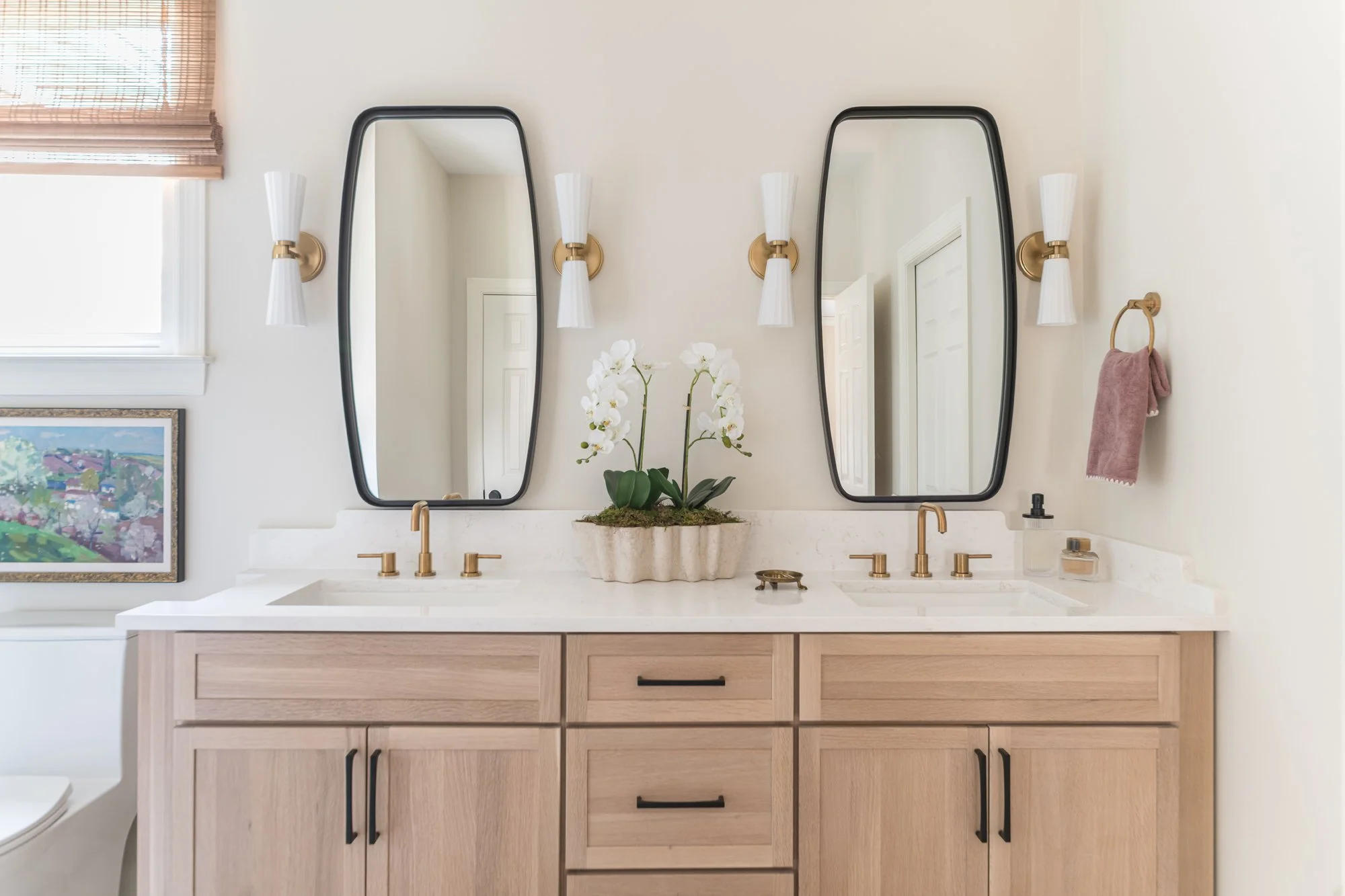 Bathroom vanity with two mirrors, floral decor, gold fixtures, vanity lights, and a pink towel hanger.