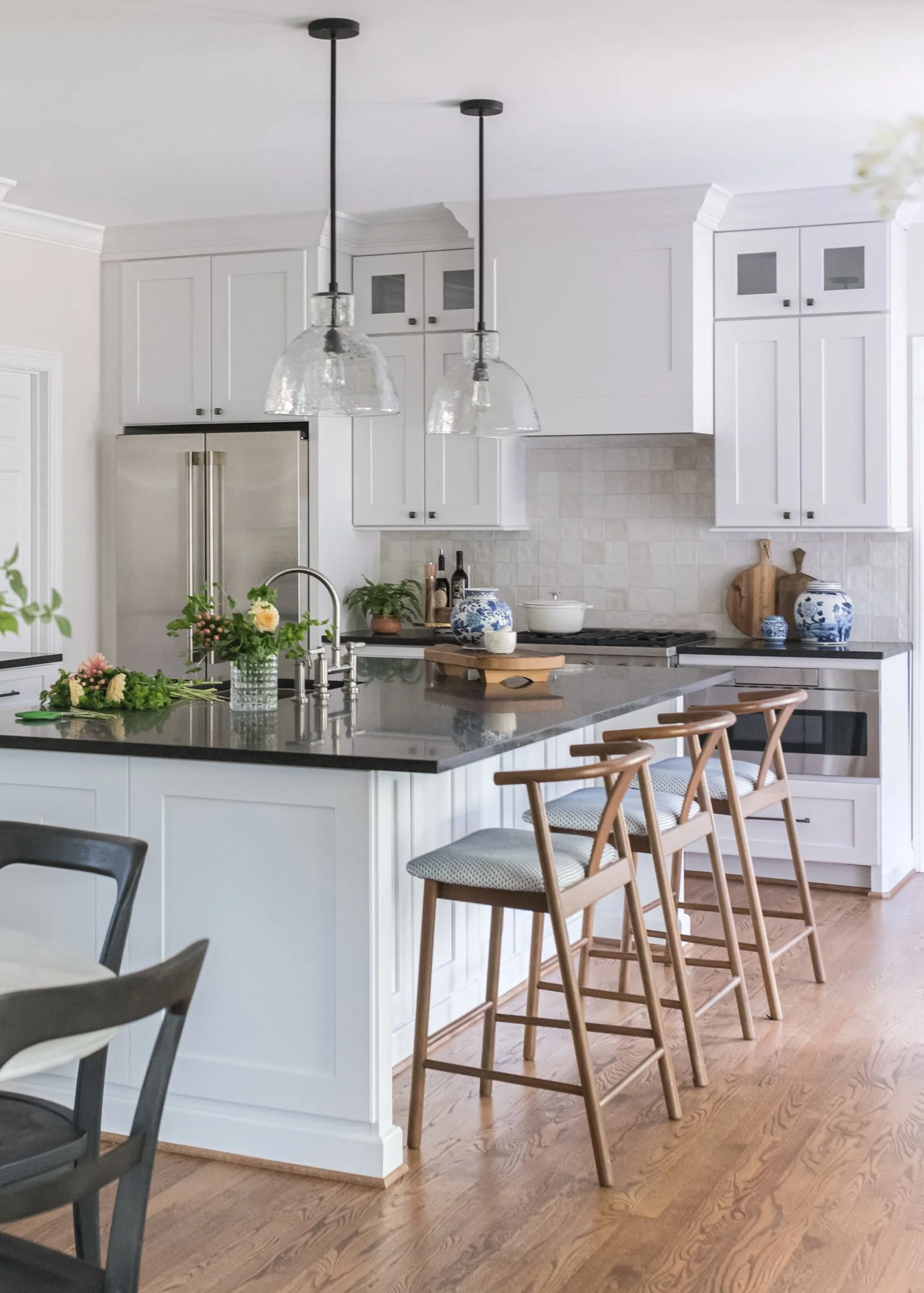Modern kitchen with white cabinets, a large kitchen island with a dark countertop, and wooden bar stools. Decor includes a bouquet of flowers, blue and white pottery, and cutting boards. Pendant lights hang above the island.