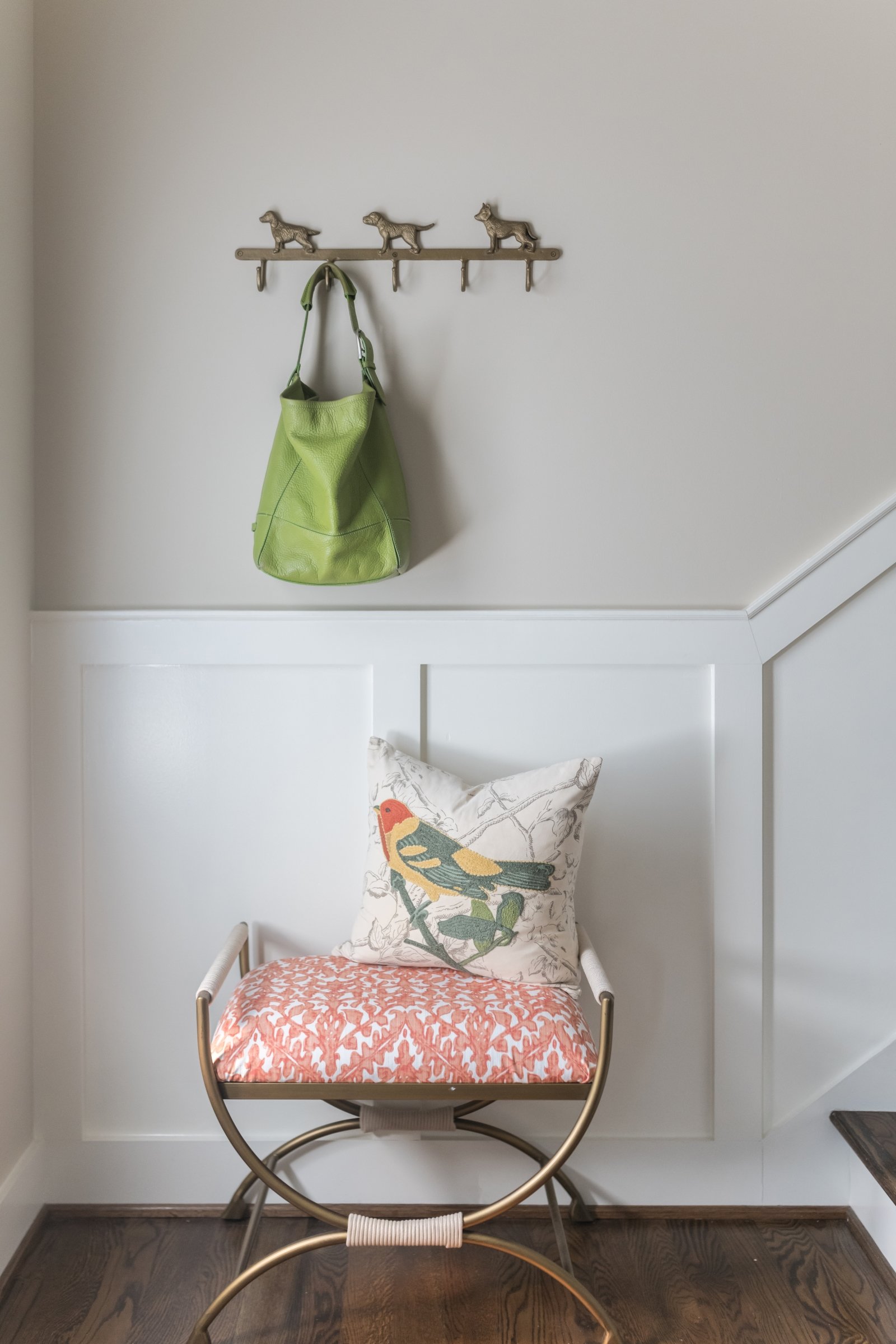 A white wall with a hook rack holding a green bag, and a chair with a pillow with a bird design and a red patterned cushion in front of the wall.