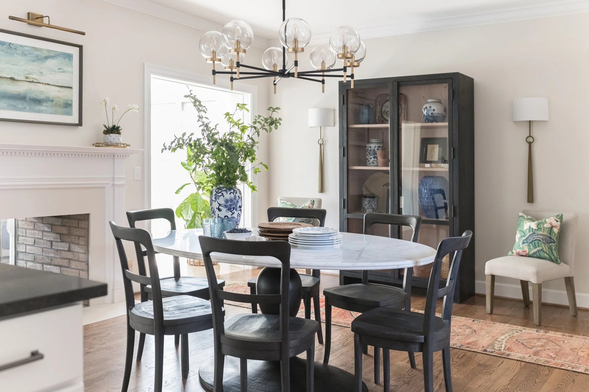 A dining room with a white marble-top table surrounded by eight black chairs. There is a large blue and white porcelain vase with greenery, a picture of seascape on the wall, a black cabinet with glass doors containing ceramic vases and pictures, and