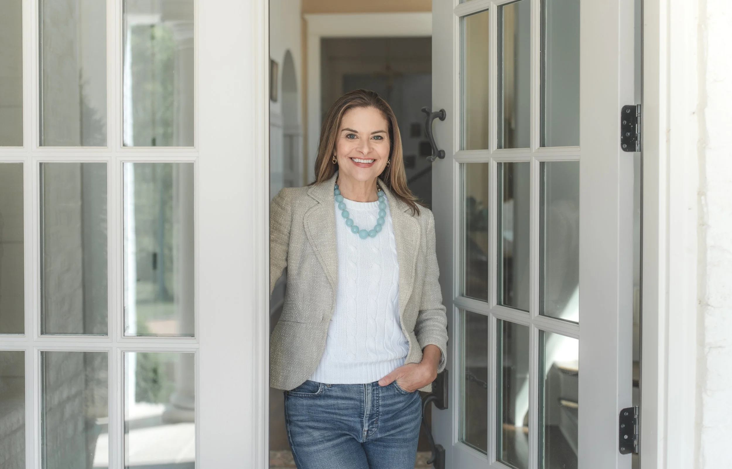 A woman smiling while standing in a doorway, wearing a light gray blazer, white sweater, blue necklace, and jeans.