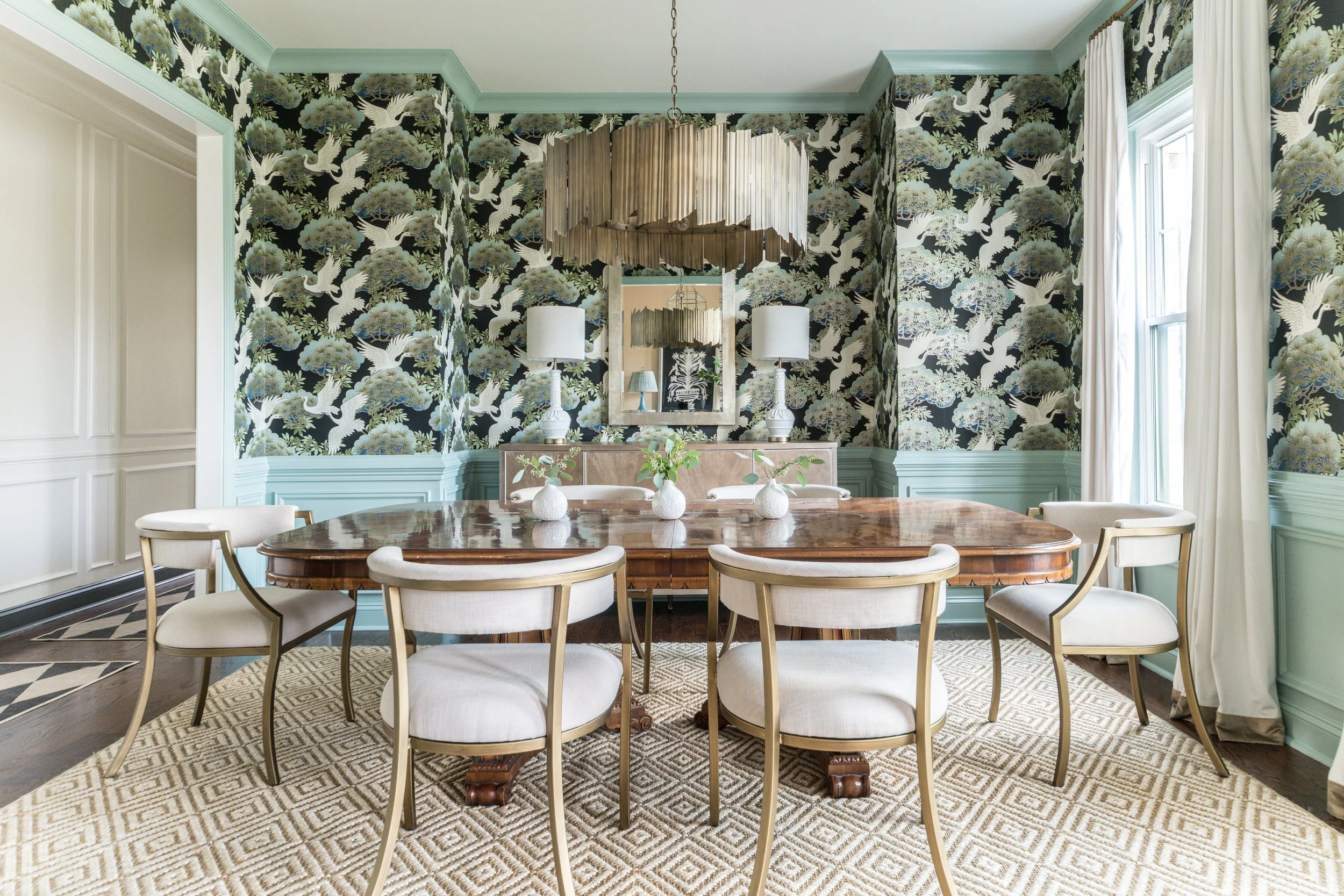 A dining room with a wooden table surrounded by beige chairs. The walls are decorated with dark floral wallpaper featuring cranes and trees. There are two white lamps on a sideboard and a rectangular mirror above it. The room has a large window with 