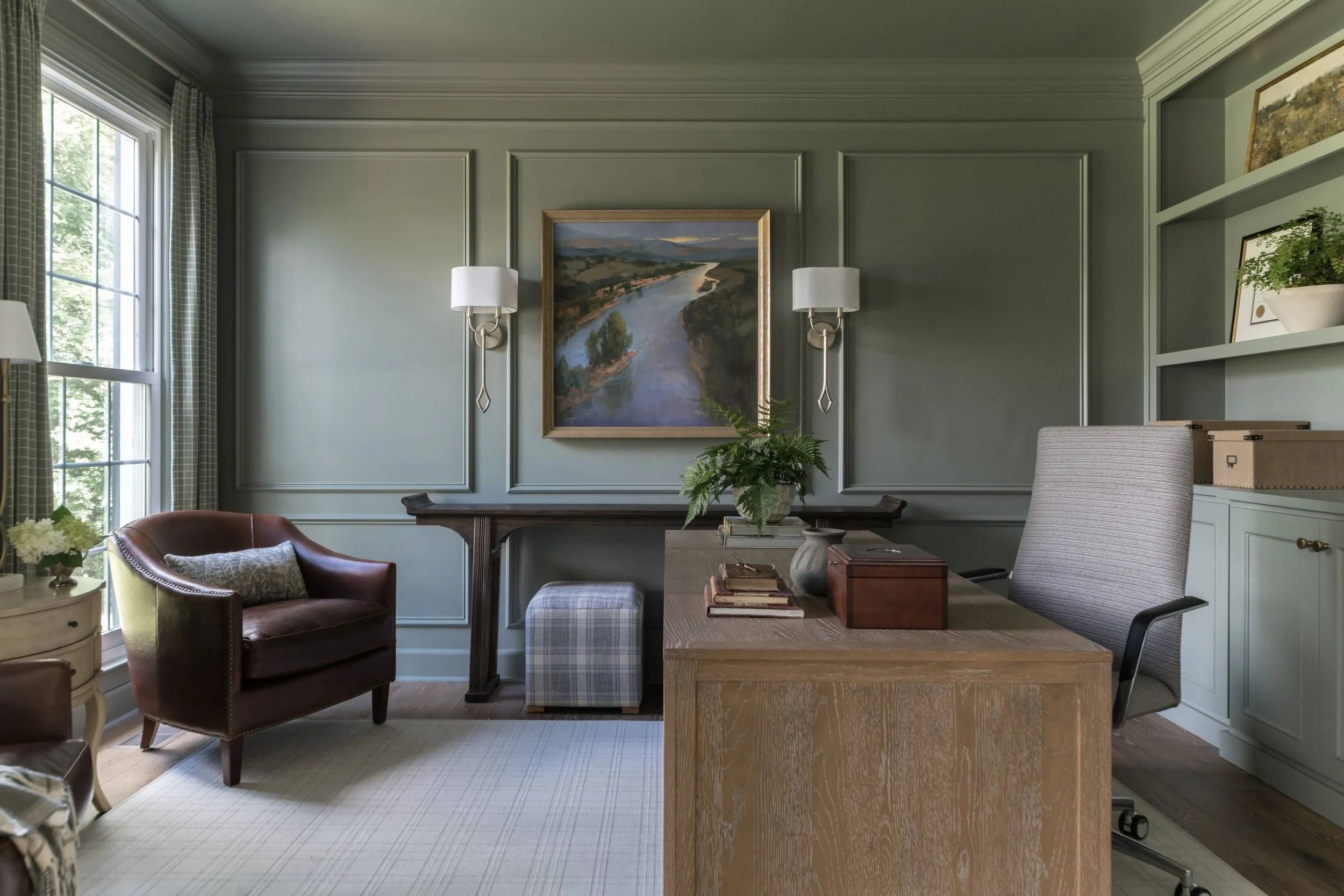 A cozy home office with green walls, a window with patterned curtains, a wooden desk with a potted fern, and assorted books and decor; a leather armchair with a pillow, and framed landscape paintings on the walls.