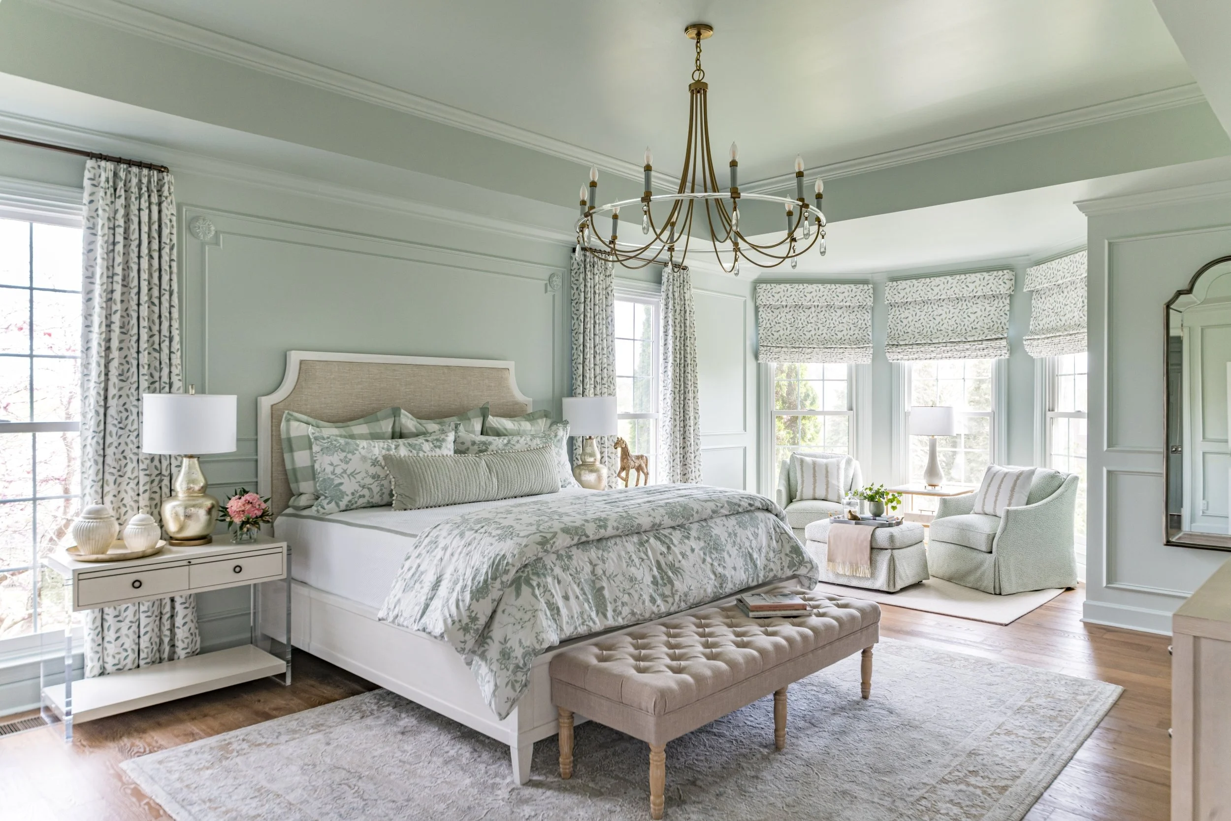 Bright bedroom with a large bed, decorative pillows, and floral bedding. There is a tufted bench at the foot of the bed, a side table with lamps, and large windows with patterned curtains and shades. A chandelier hangs from the ceiling, and cozy seat