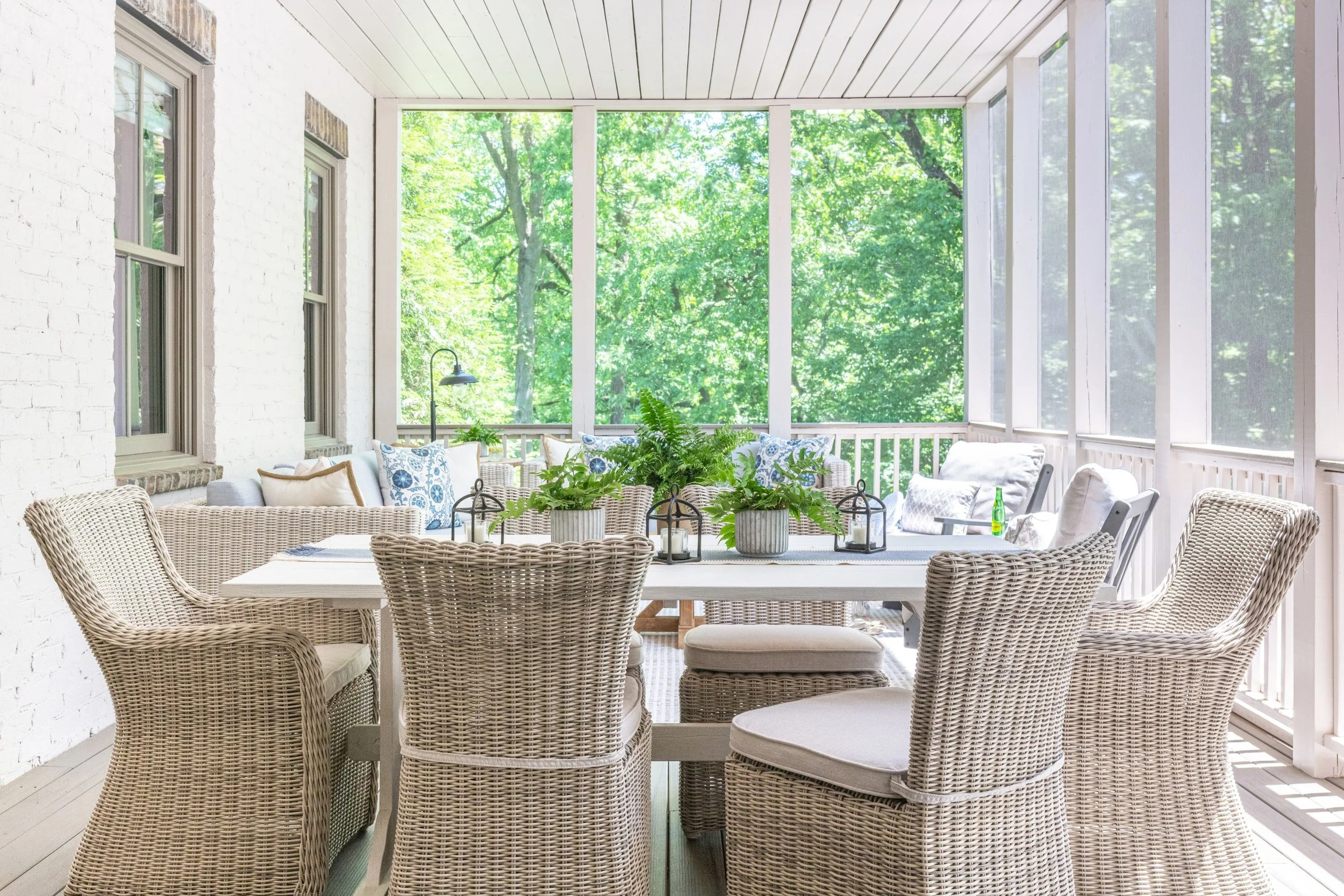 Sunlit covered porch with wicker chairs, a white dining table, and potted plants, overlooking lush green trees.