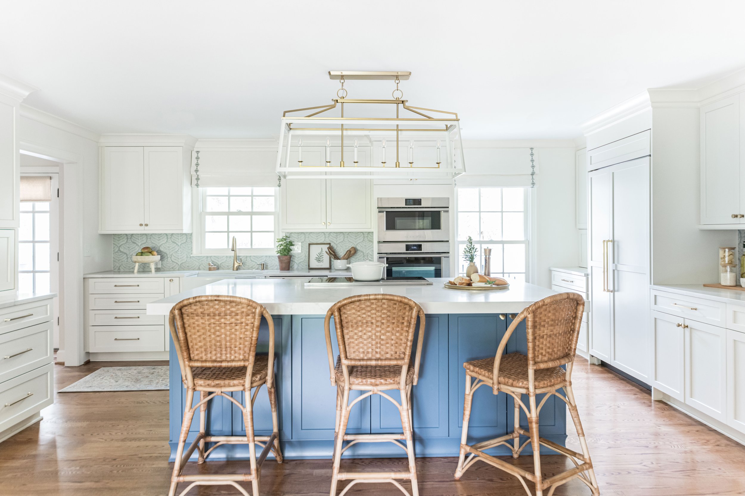 Bright, modern kitchen with white cabinets, a blue kitchen island, and natural woven chairs around a central island. Large windows with simple window coverings let in ample natural light.