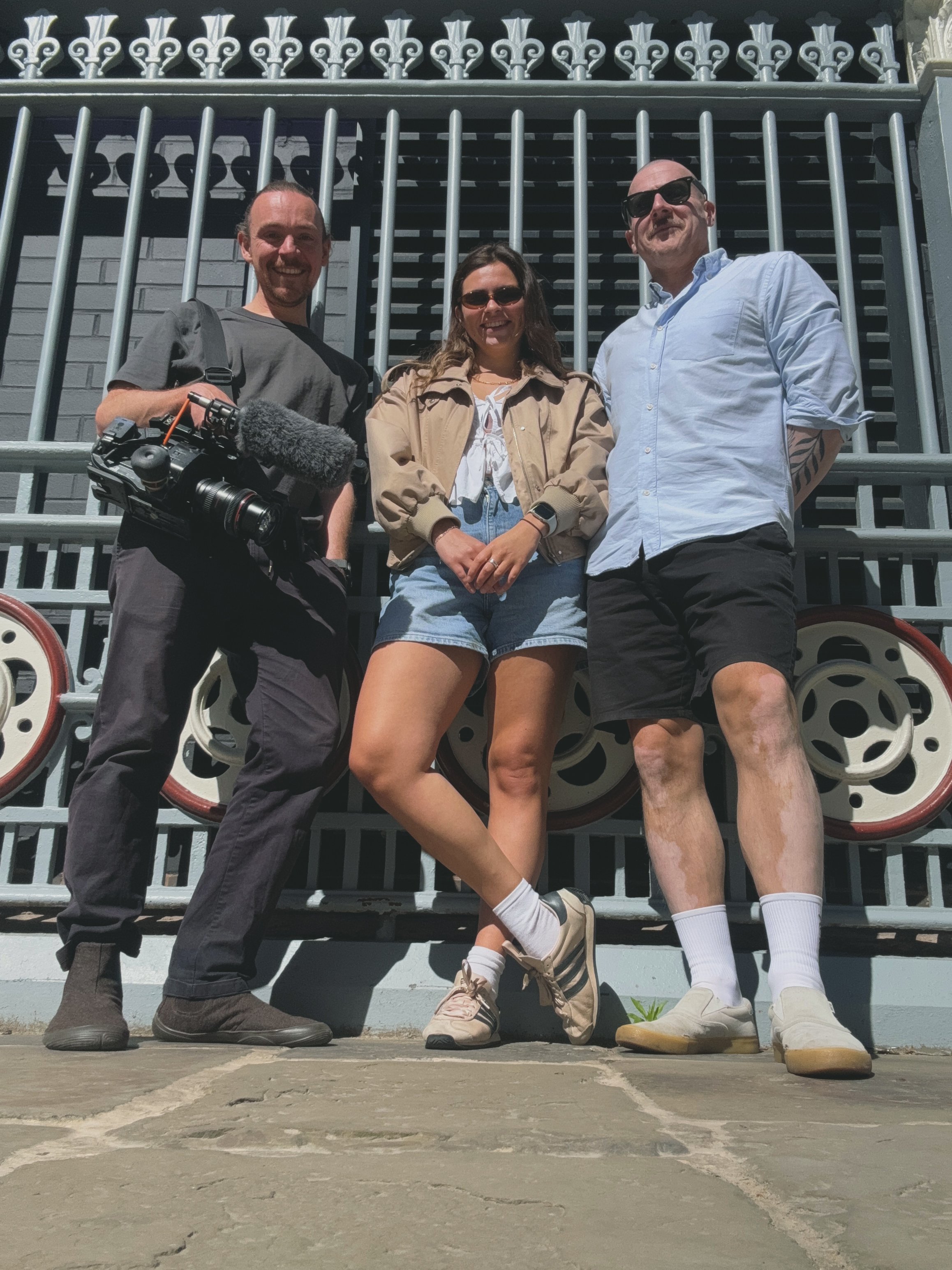 Three friends standing in front of a large metal gate, smiling at the camera. The person on the left holds a camera, the woman in the middle is wearing sunglasses, a beige jacket, and jean shorts, and the man on the right is wearing sunglasses, a light blue shirt, and shorts. They are outdoors on a sunny day.
