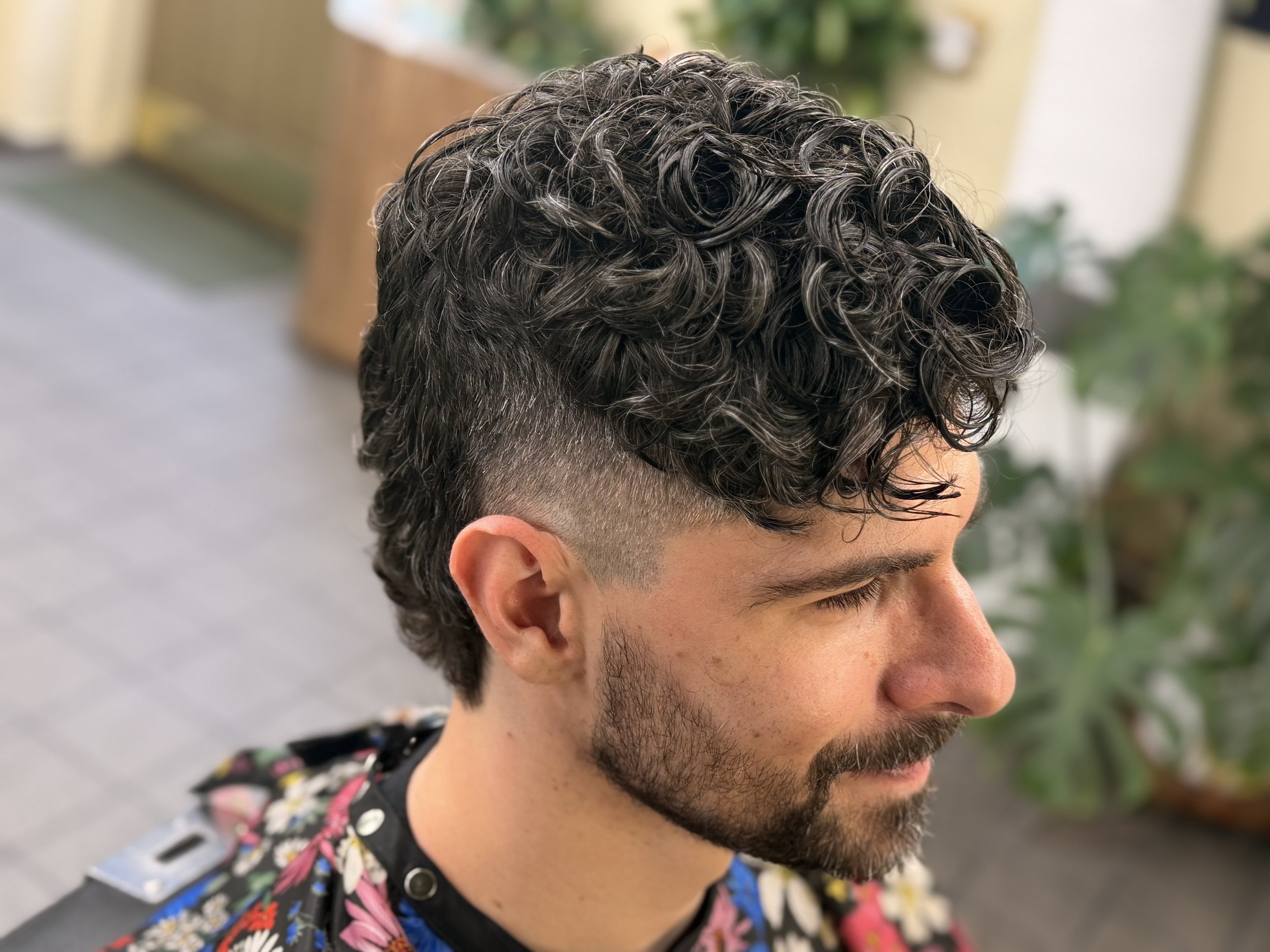 Side view of a man with dark, curly hair and a beard, wearing a floral shirt, outdoors.